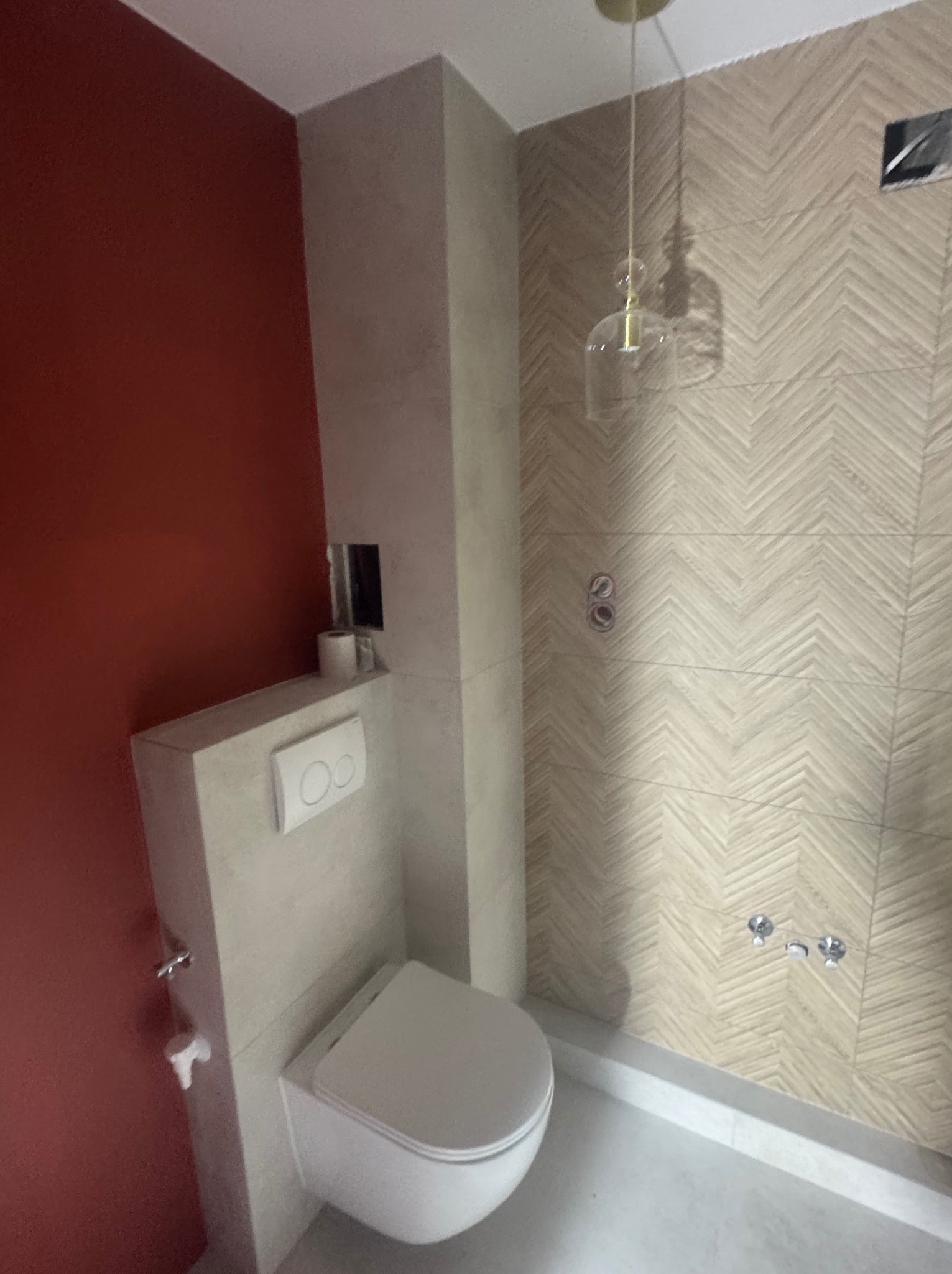 Nowoczesna łazienka po remoncie: podwieszana toaleta, ściana z wzorem jodełki, czerwona ściana, minimalistyczna lampa. Widok na fragment wykończonego wnętrza.