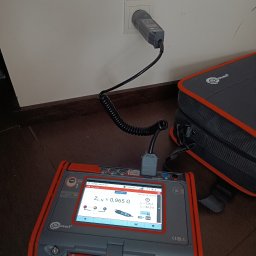 KOZIOŁ ELECTRIC ŁUKASZ KOZIOŁ - Miernik impedancji pętli zwarcia Sonel podłączony do gniazdka elektrycznego, wyświetlający wynik pomiaru ZL-N = 0,965 Ω, obok torba narzędziowa Sonel.