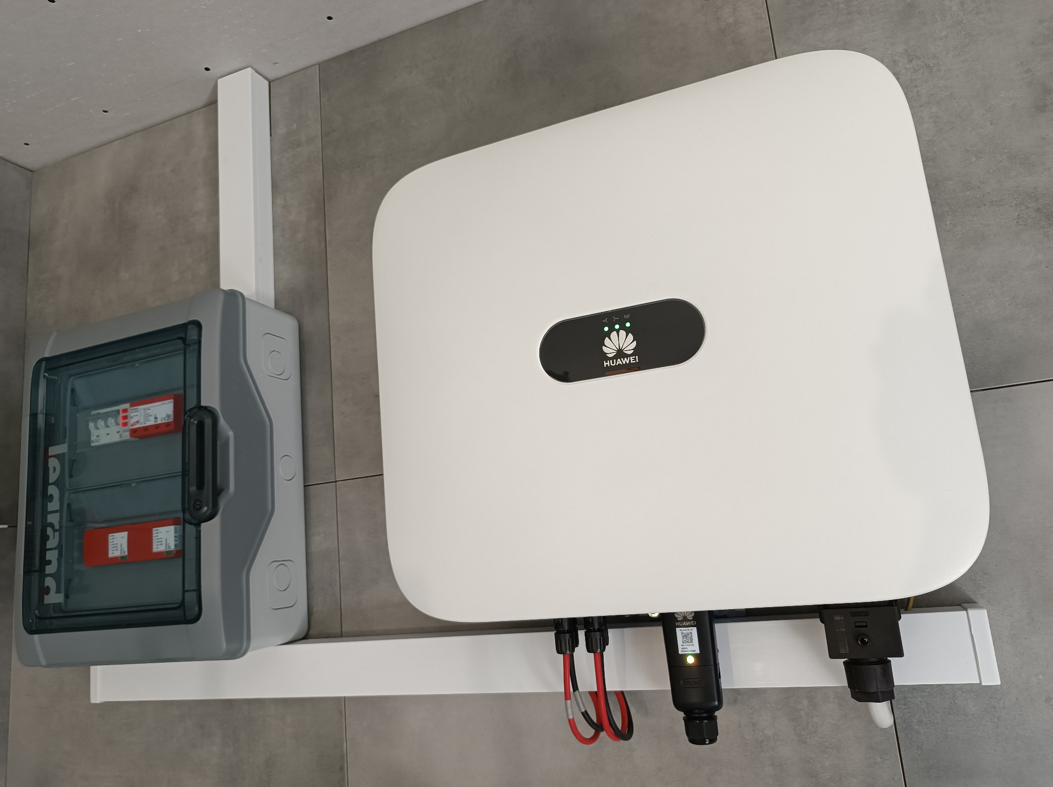 Szara skrzynka elektryczna Legrand i biały falownik Huawei zamontowane na ścianie z szarych płytek, z widocznymi przewodami.