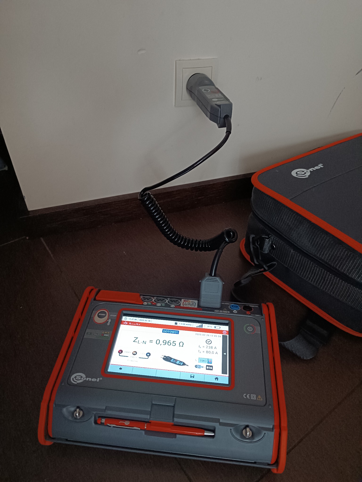 Miernik impedancji pętli zwarcia Sonel podłączony do gniazdka elektrycznego, wyświetlający wynik pomiaru ZL-N = 0,965 Ω, obok torba narzędziowa Sonel.