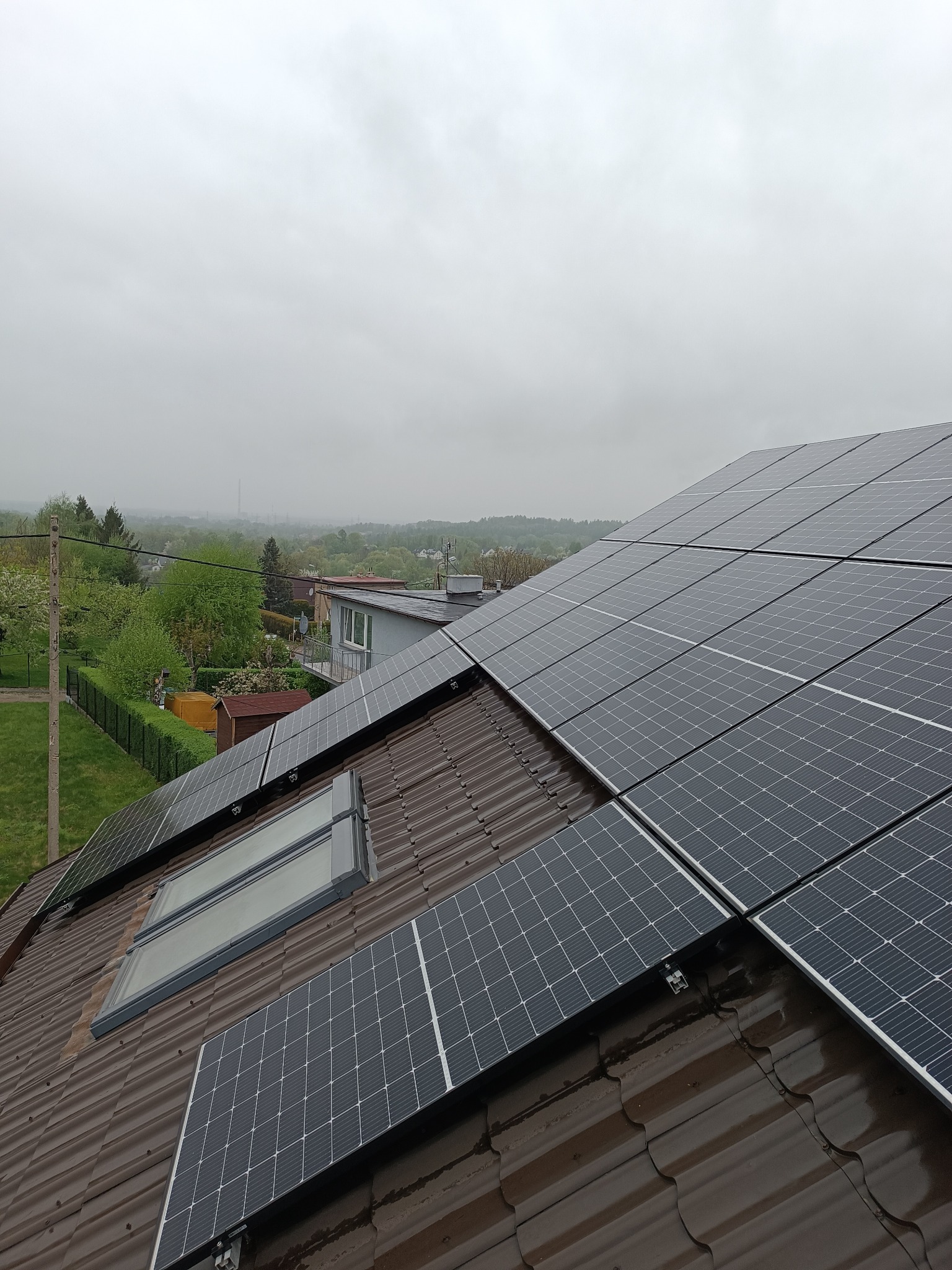Panel słoneczny na brązowym dachu z blachodachówki, widoczne okna dachowe i odległy krajobraz w pochmurny dzień.