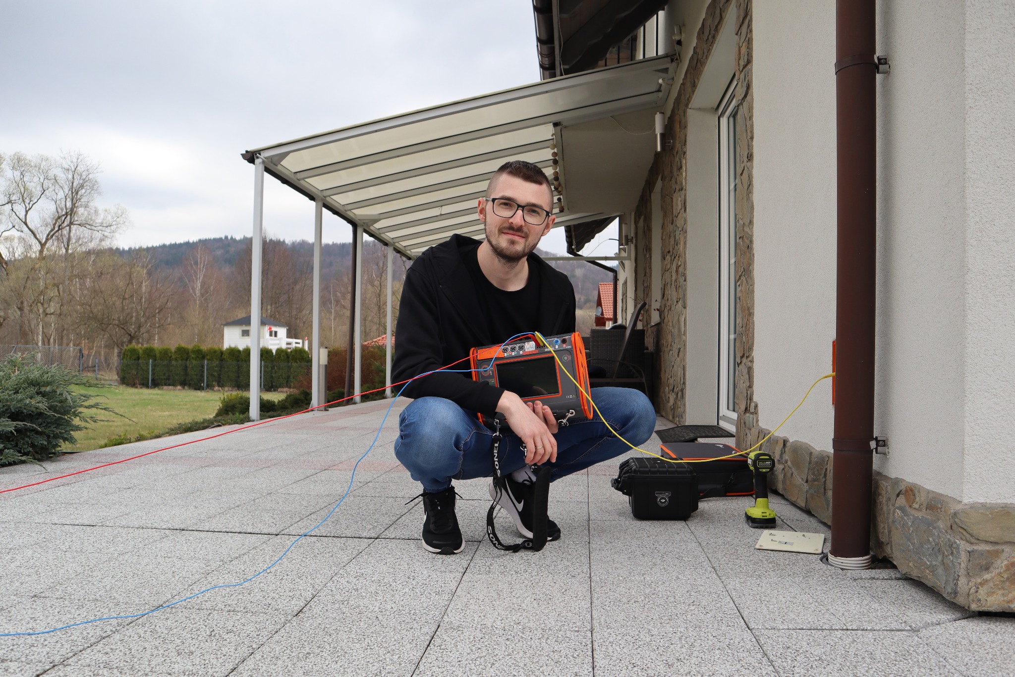 Mężczyzna w okularach wykonuje pomiary elektryczne przy budynku, używając specjalistycznego pomarańczowego miernika z podłączonymi przewodami.