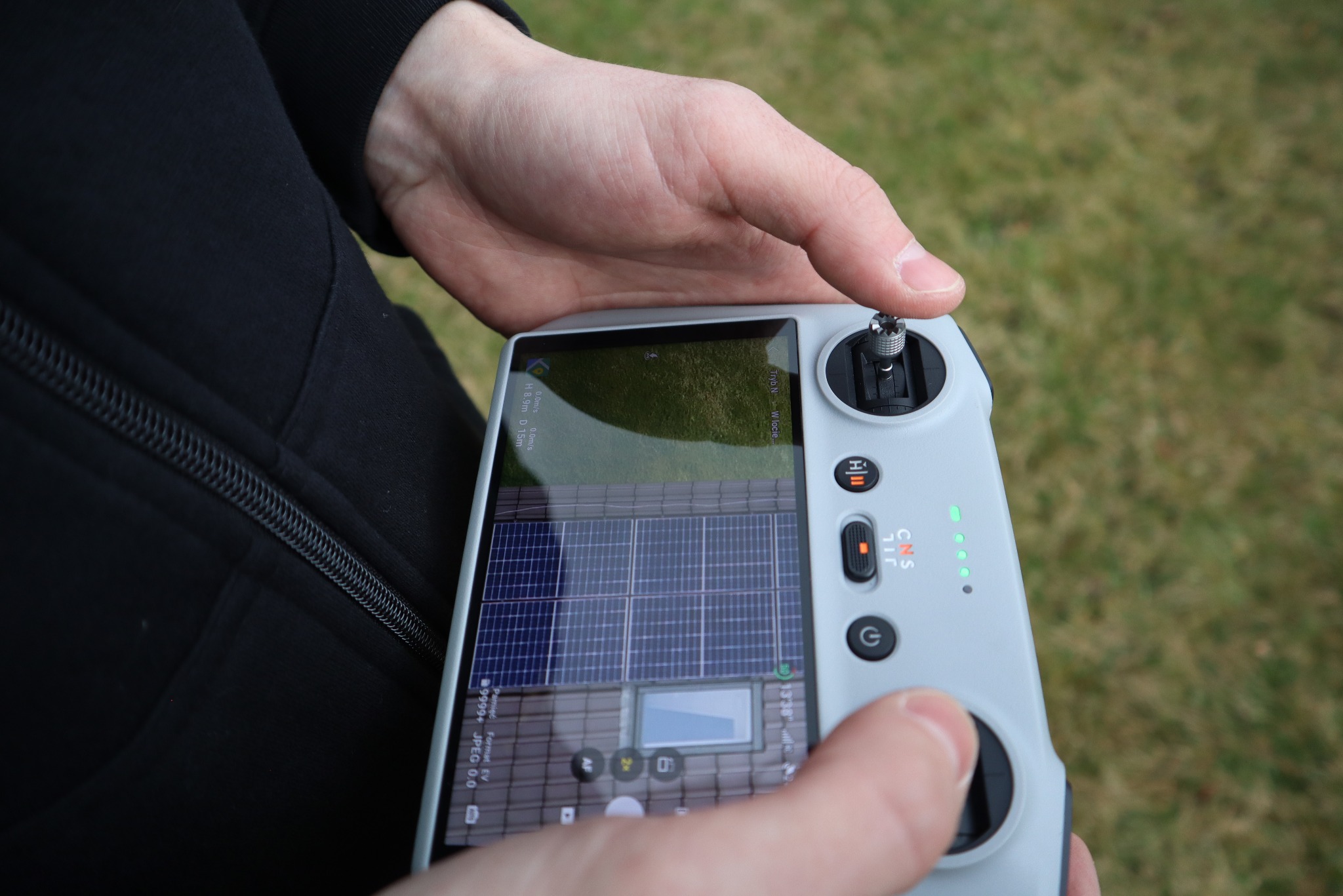 Operator drona, trzymając kontroler, filmuje panele słoneczne na dachu. Widoczny ekran kontrolera z podglądem na żywo z kamery drona.