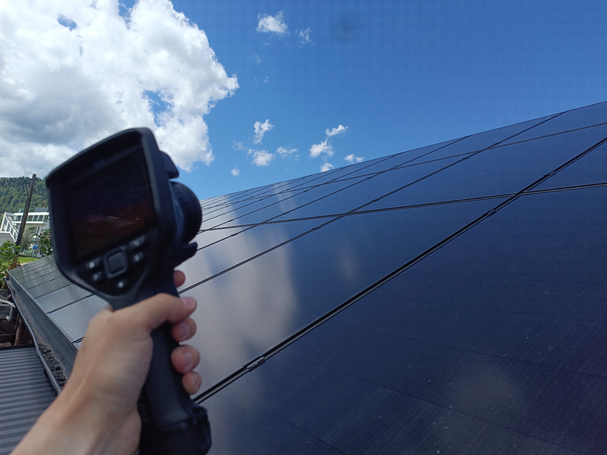 Termowizja paneli słonecznych na dachu, wykonywana z bliska przy słonecznej pogodzie. Widoczna ręka trzymająca kamerę termowizyjną.