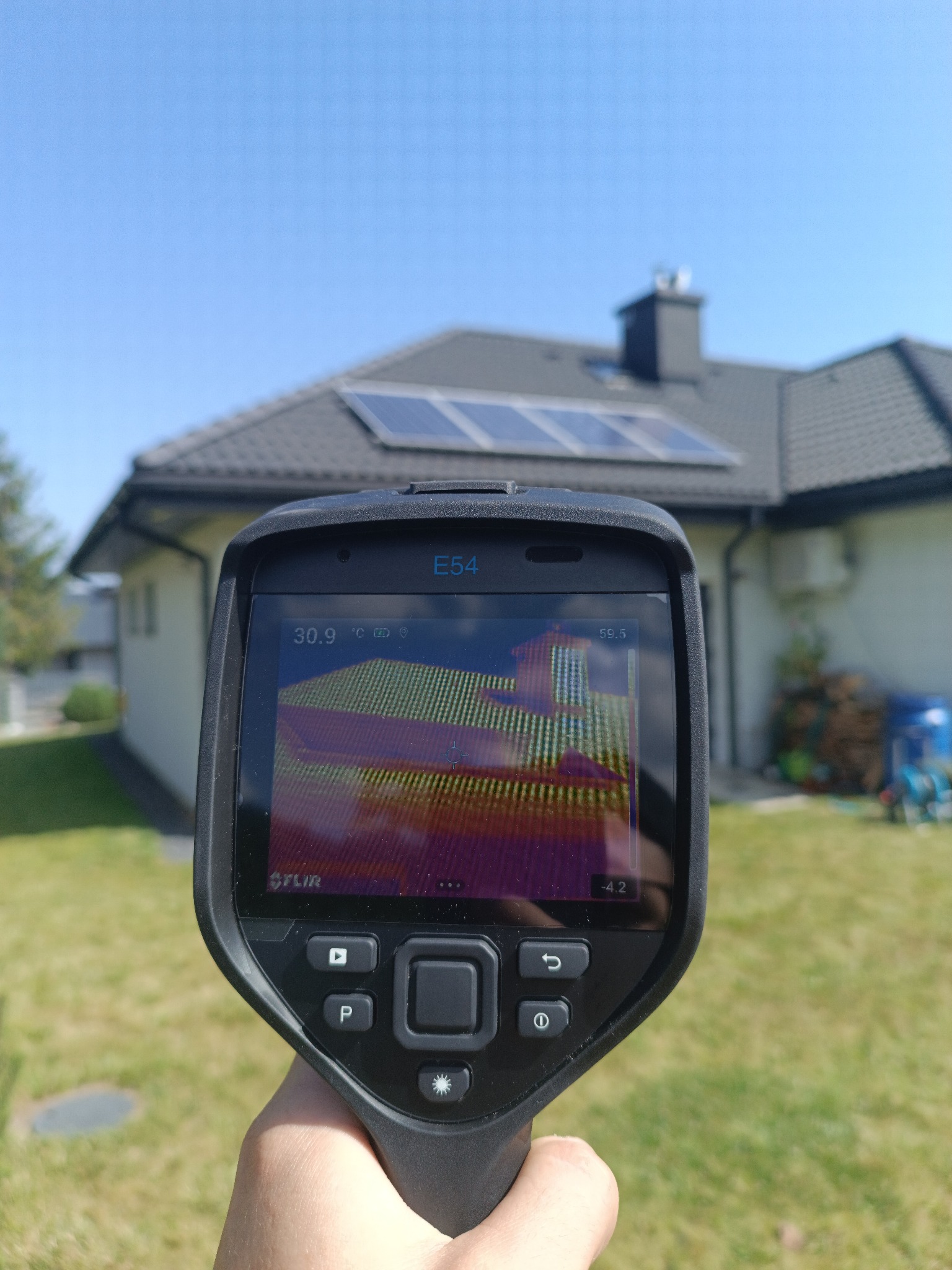 Termowizja domu jednorodzinnego z panelami słonecznymi, uchwycona z bliska urządzeniem FLIR E54. Widoczny ekran z rozkładem temperatur na elewacji budynku.