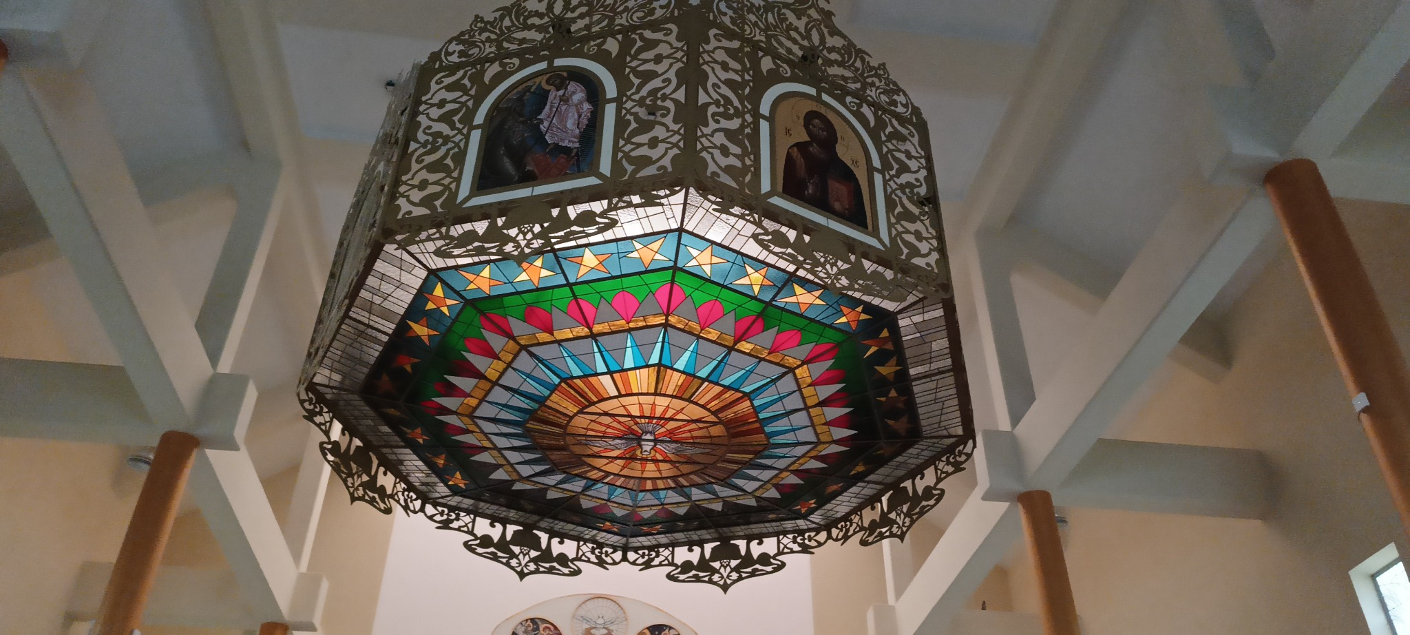 Ozdobny, metalowy żyrandol z witrażowym wypełnieniem i motywami religijnymi, zawieszony w kościele lub kaplicy, widziany z dołu.