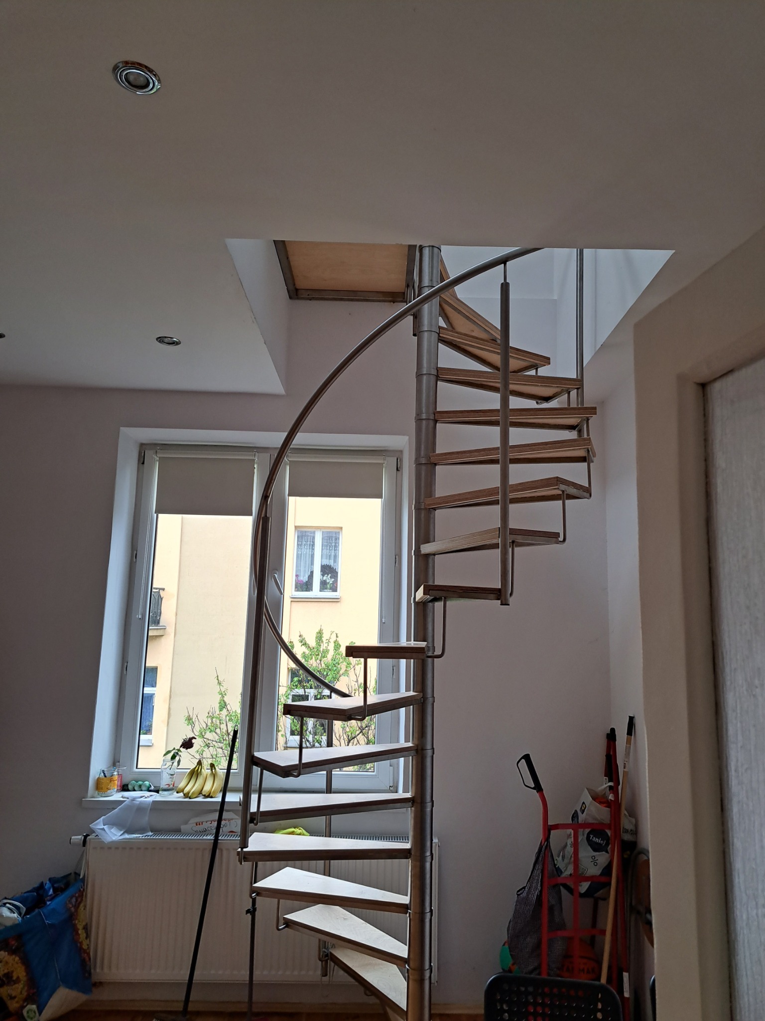 Spiralne schody z drewnianymi stopniami i metalową konstrukcją w jasnym wnętrzu, widok na okno z parapetem i fragment wyposażenia.