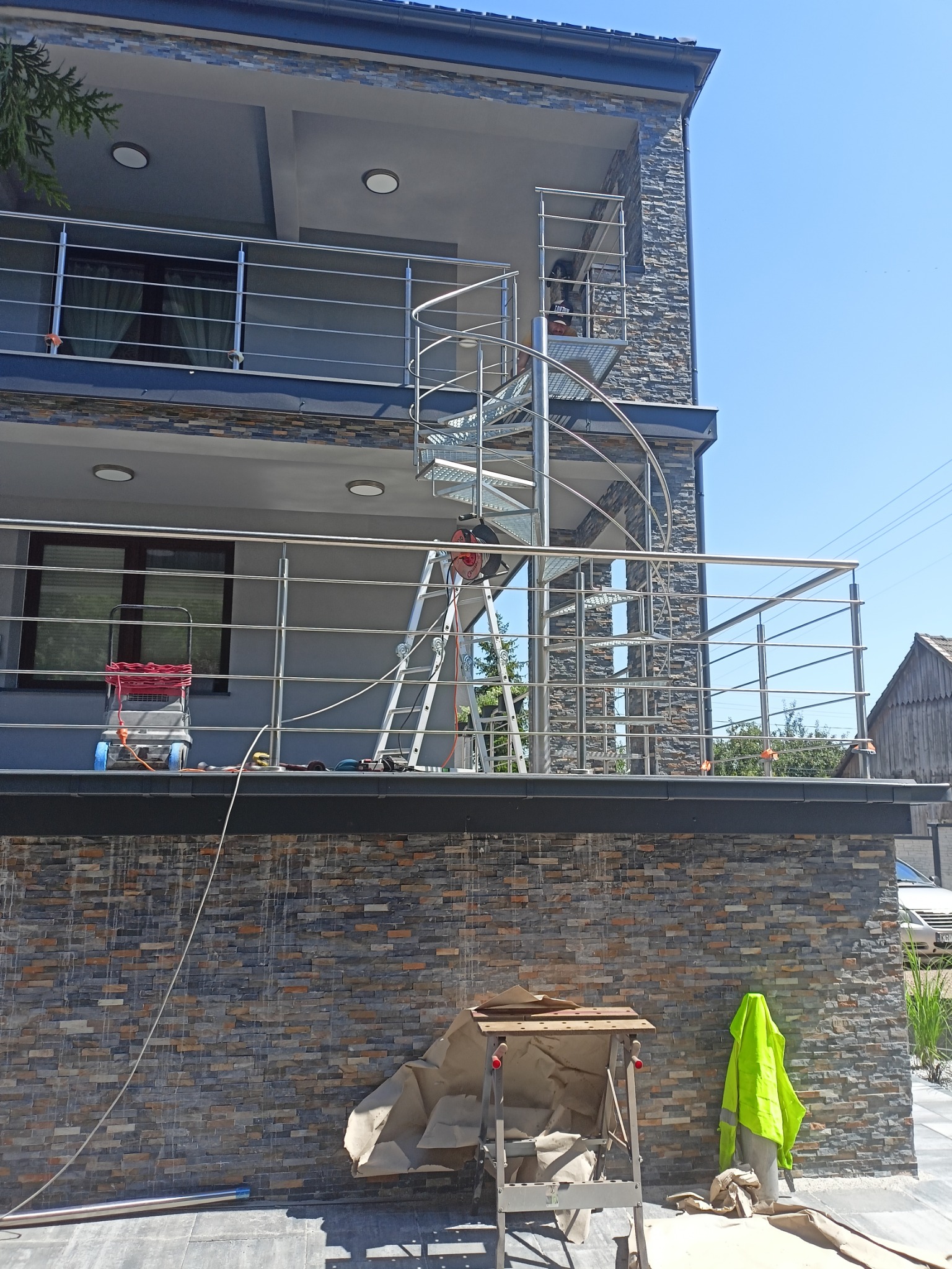 Montaż zewnętrznej balustrady nierdzewnej na balkonie z spiralnymi schodami, widoczny pracownik na schodach i narzędzia pracy.