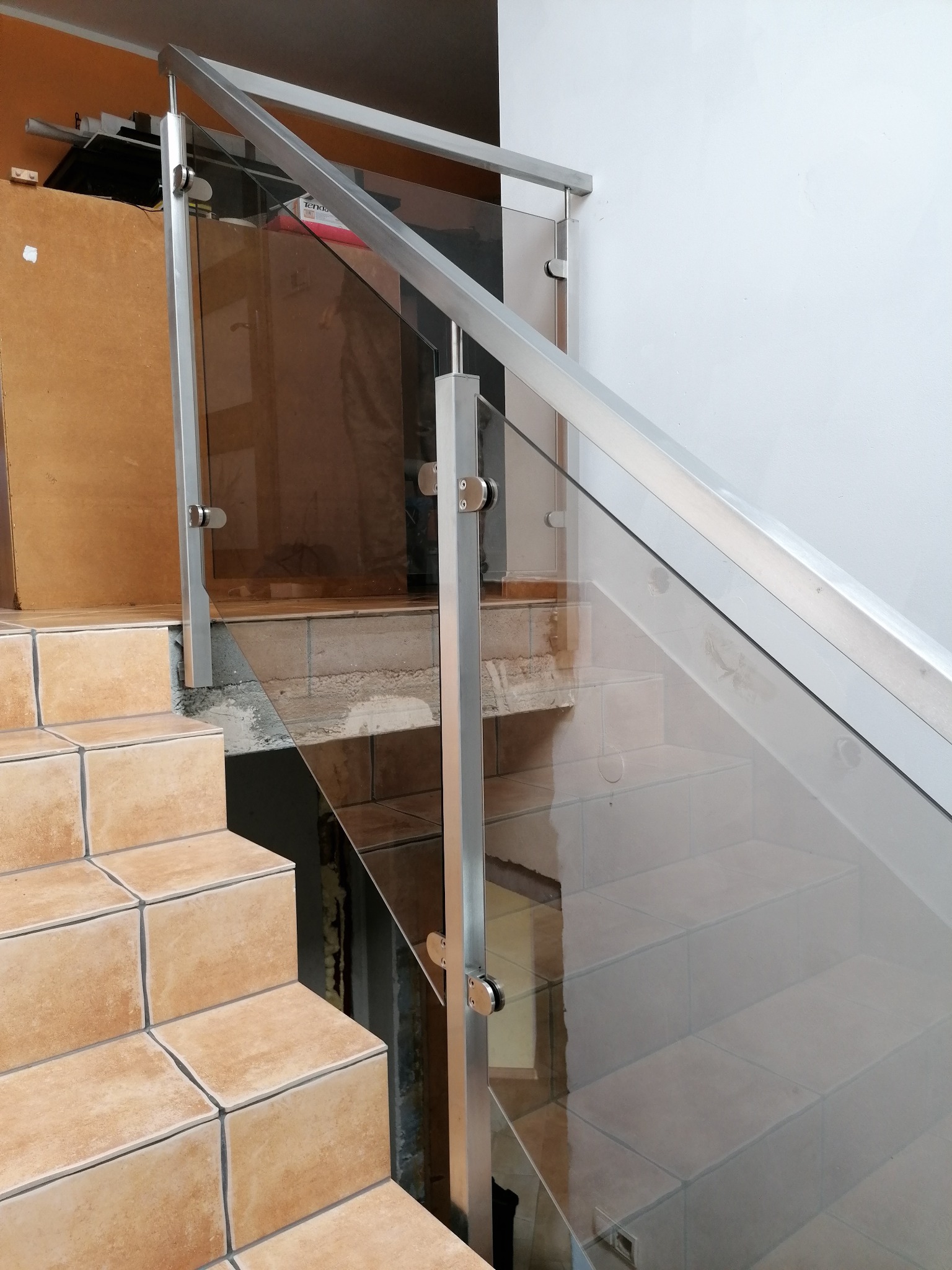 Szklana balustrada z metalowymi słupkami zamocowana na schodach wyłożonych terakotą, widok z dołu na górę.