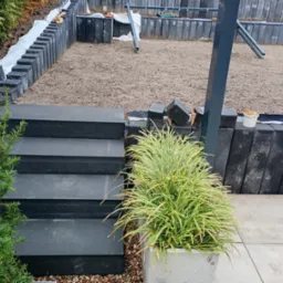 Budowa tarasu z czarnych, betonowych elementów: schody, murki oporowe z izolacją, przygotowane podłoże, widoczny fragment konstrukcji zadaszenia i donica z trawą ozdobną.