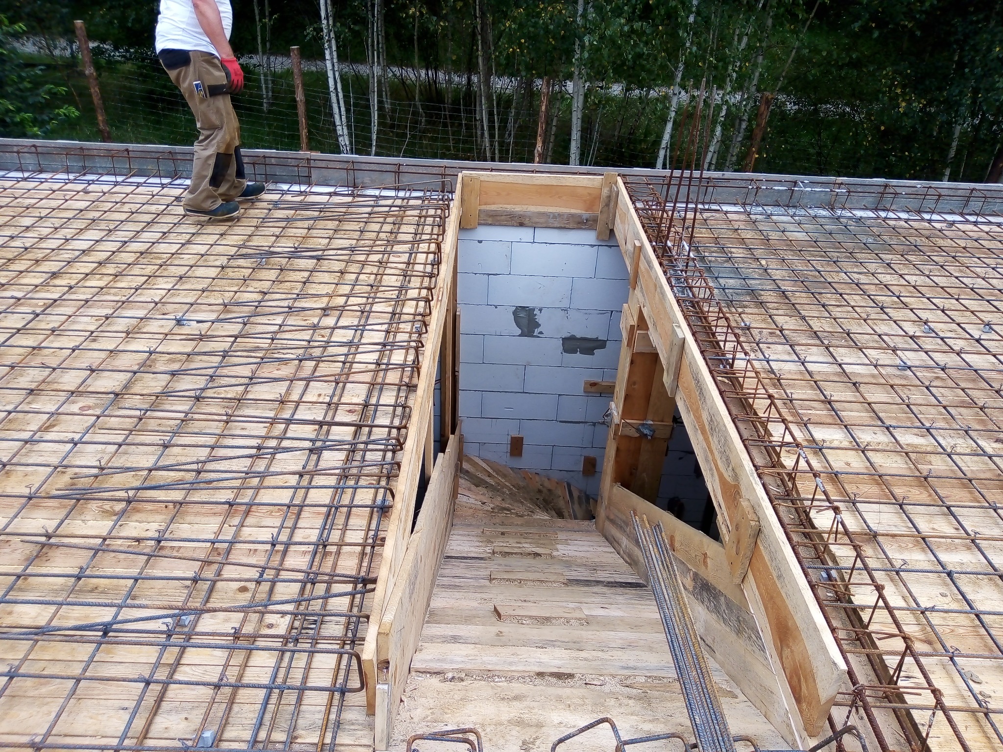 Zbrojenie stropu z otworem na schody, widok z góry na drewniany szalunek i siatkę z prętów stalowych, osoba w odzieży roboczej w tle.