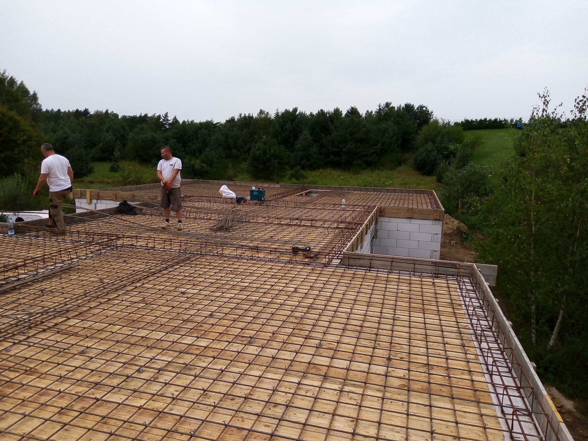 Zbrojenie płyty fundamentowej na drewnianym szalunku z widocznymi pracownikami i narzędziami na tle zieleni.