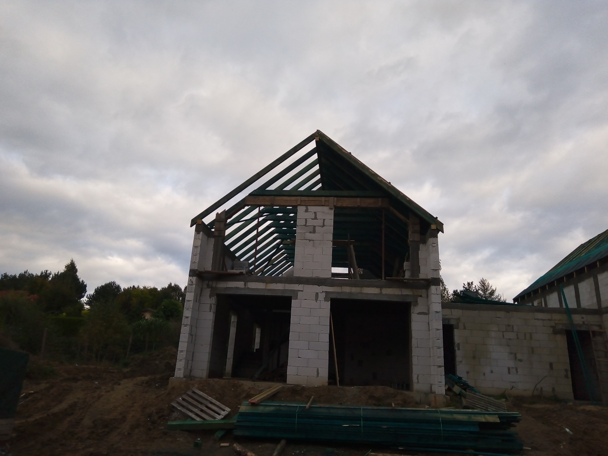 Surowy stan dwukondygnacyjnego domu z białych bloczków, widoczne drewniane elementy konstrukcji dachu, na pierwszym planie ułożone zielone profile, pochmurne niebo w tle.