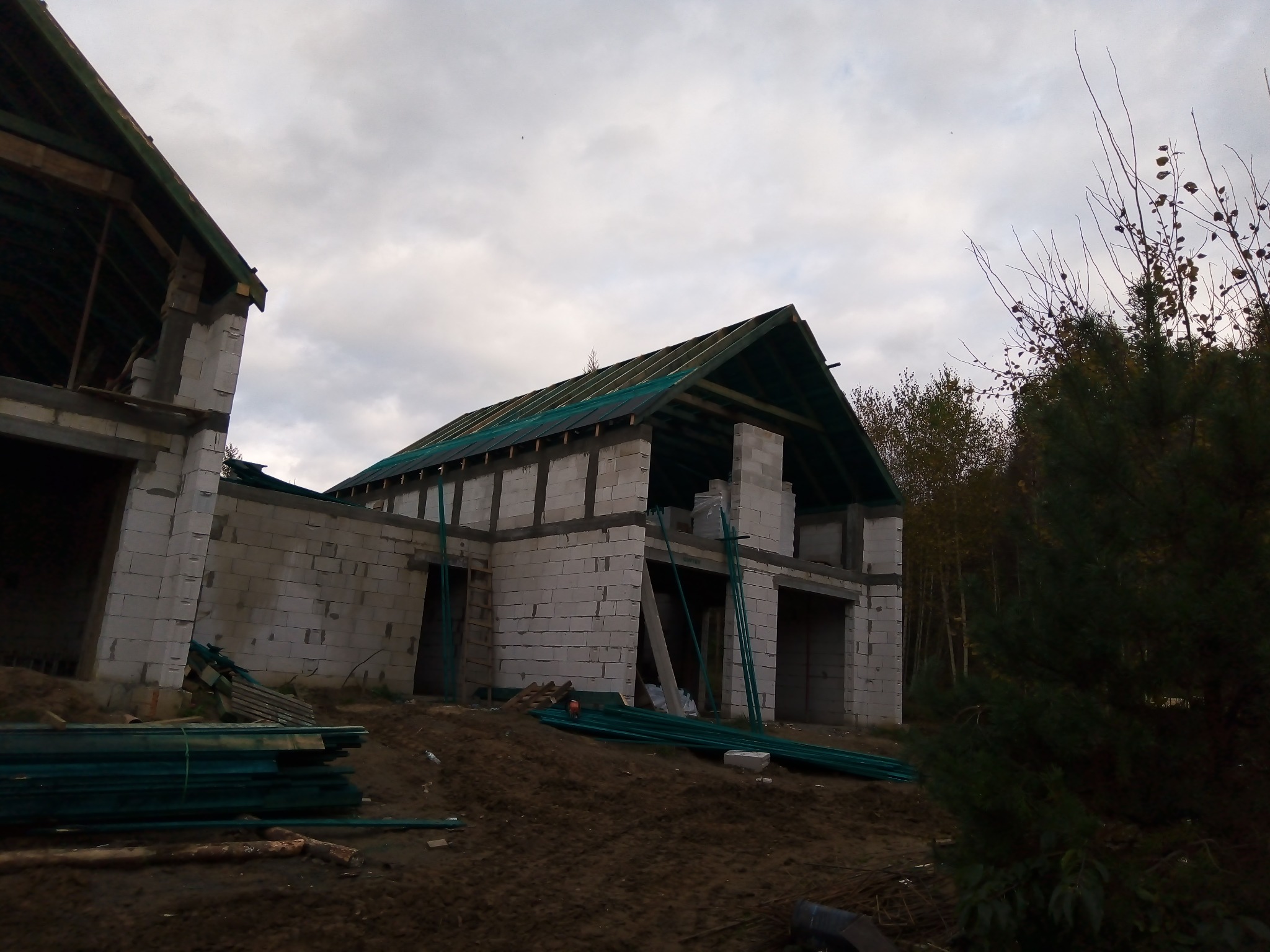 Surowy stan budowy domu jednorodzinnego z białych bloczków, widoczne drewniane elementy konstrukcji dachu i zielone rury na ziemi, pochmurne niebo w tle.