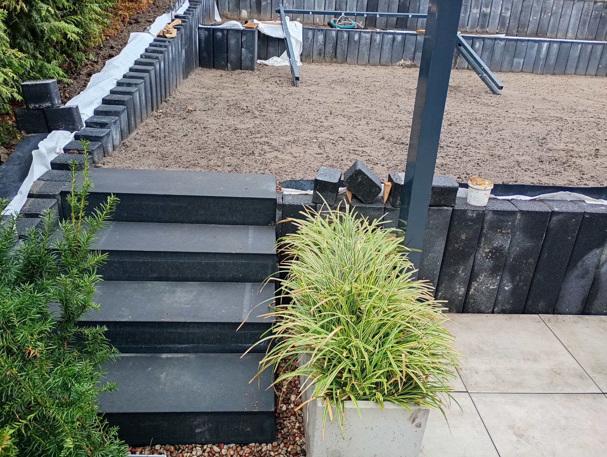 Budowa tarasu z czarnych, betonowych elementów: schody, murki oporowe z izolacją, przygotowane podłoże, widoczny fragment konstrukcji zadaszenia i donica z trawą ozdobną.