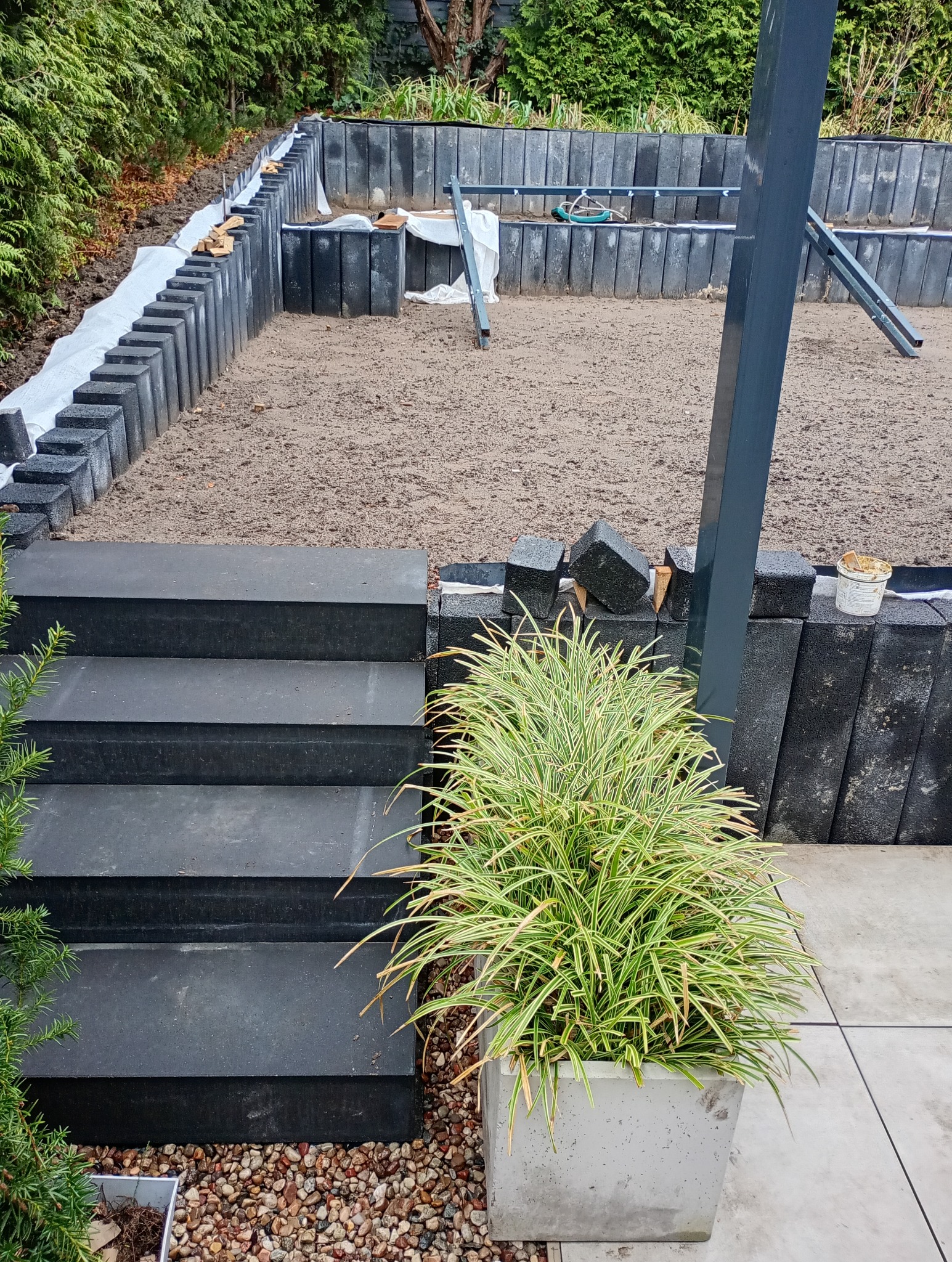 Budowa tarasu z czarnych bloczków betonowych, widoczne schody, rabata w trakcie tworzenia, ziemia przygotowana pod nasadzenia, drabina w tle, roślina ozdobna w donicy na pierwszym planie.