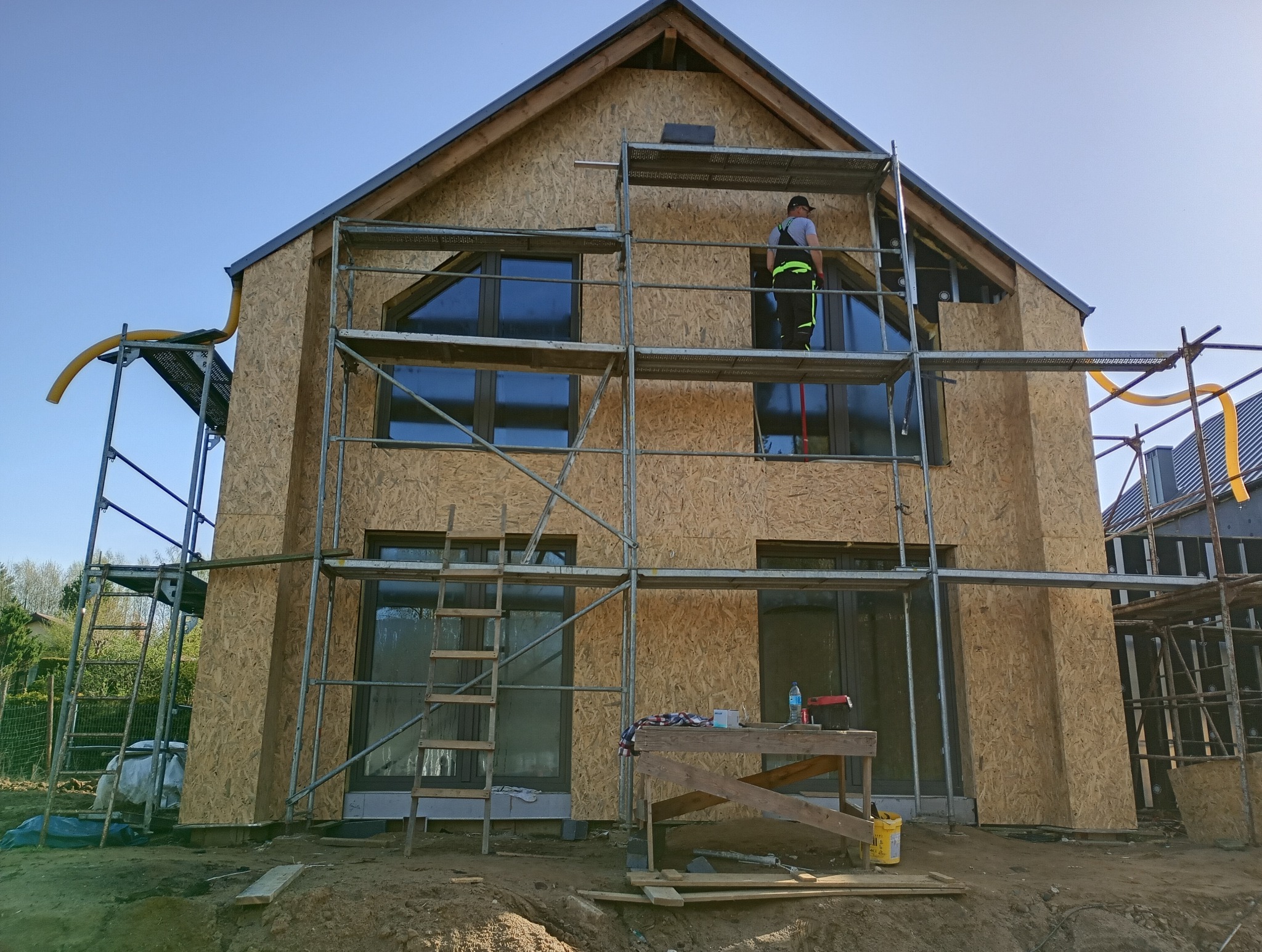 Dom w trakcie budowy, obłożony płytami OSB, widoczne rusztowania i pracownik na wysokości, żółty wąż wentylacyjny przy dachu.