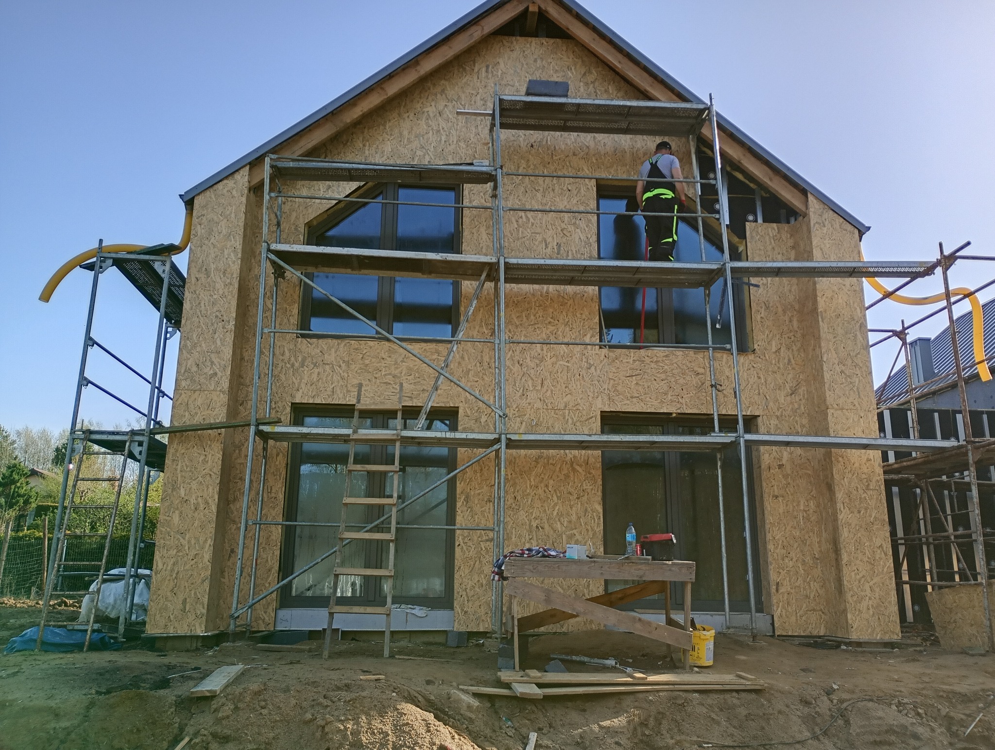 Budowa domu jednorodzinnego: robotnik na rusztowaniu montuje płyty OSB na elewacji, widoczne okna, żółty wąż wentylacyjny i narzędzia na prowizorycznym stole.