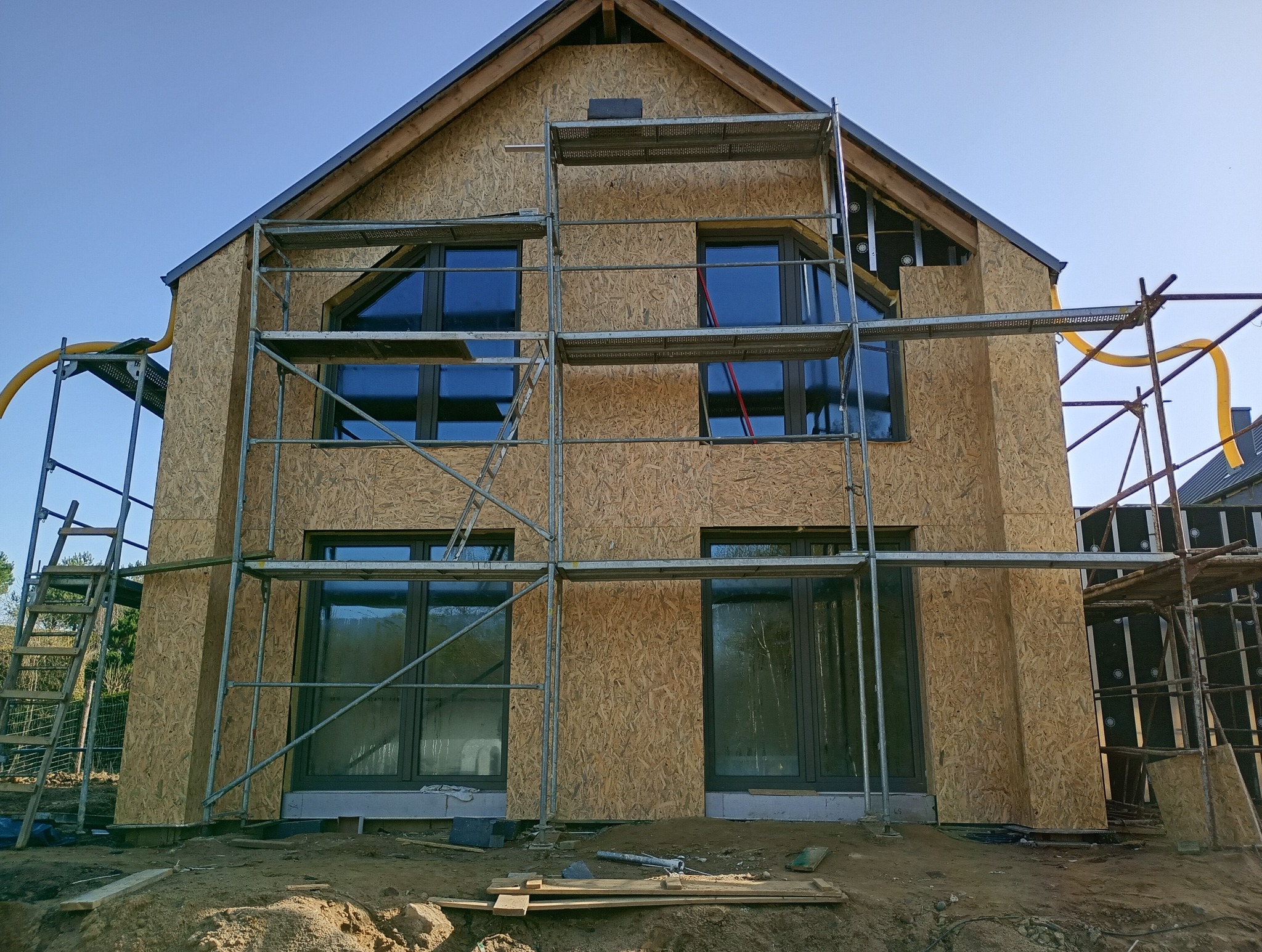 Budowa domu w stanie surowym zamkniętym: ściany obłożone płytami OSB, okna wstawione, rusztowania wokół budynku, widoczny fragment instalacji wentylacyjnej.