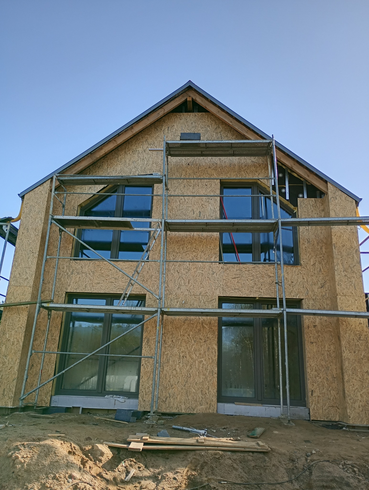 Budynek w trakcie budowy, widoczne płyty OSB na elewacji, okna z ciemnymi ramami oraz rusztowanie, ziemia wokół budynku.
