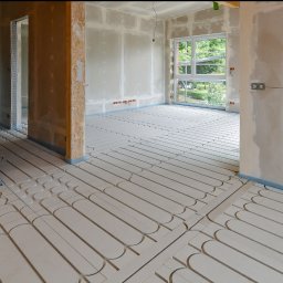 PERFECT - Surowe wnętrze w trakcie prac wykończeniowych: widoczne ogrzewanie podłogowe, ściany przygotowane do malowania, sufit z widoczną instalacją elektryczną, okno z widokiem na zieleń.