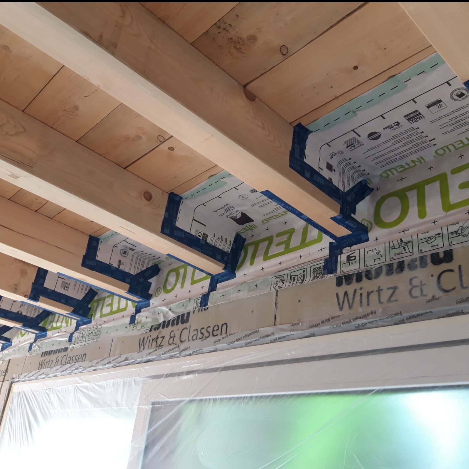 Montaż folii paroizolacyjnej z taśmą uszczelniającą na drewnianej konstrukcji stropu, widoczne logo Intello i Wirtz & Classen.