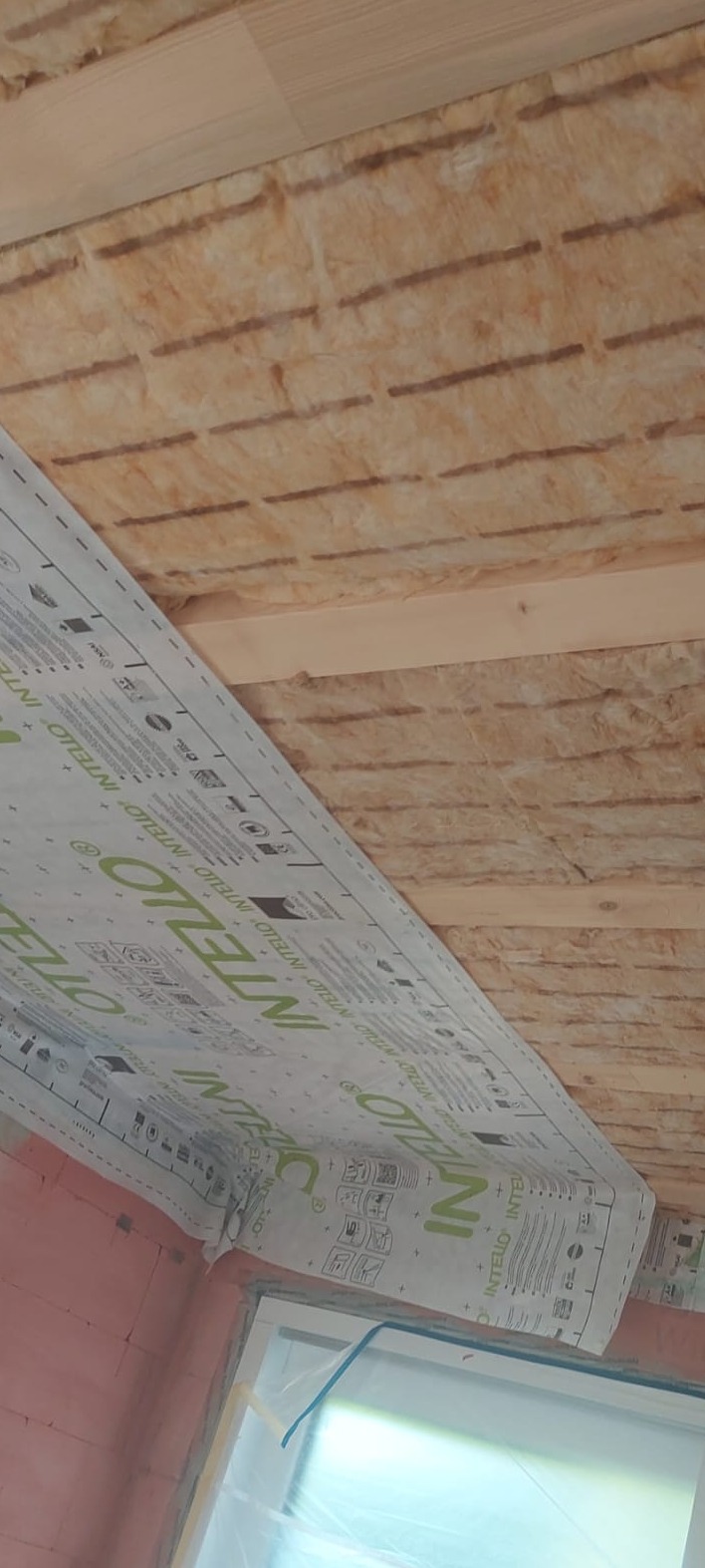 Montaż izolacji termicznej z folią paroizolacyjną na poddaszu, widoczne wełna mineralna, drewniana konstrukcja oraz okno zabezpieczone folią ochronną.