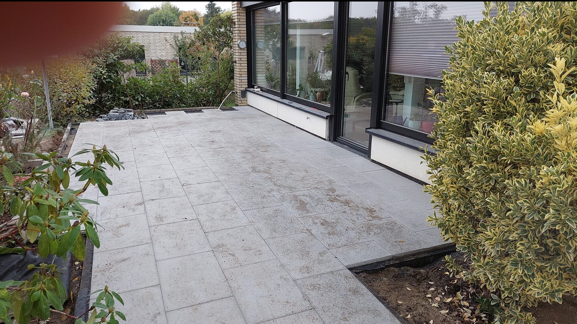Szara, kwadratowa kostka brukowa na tarasie przylegającym do domu z dużymi oknami, widoczne rośliny w ogrodzie i częściowo ułożona nawierzchnia.
