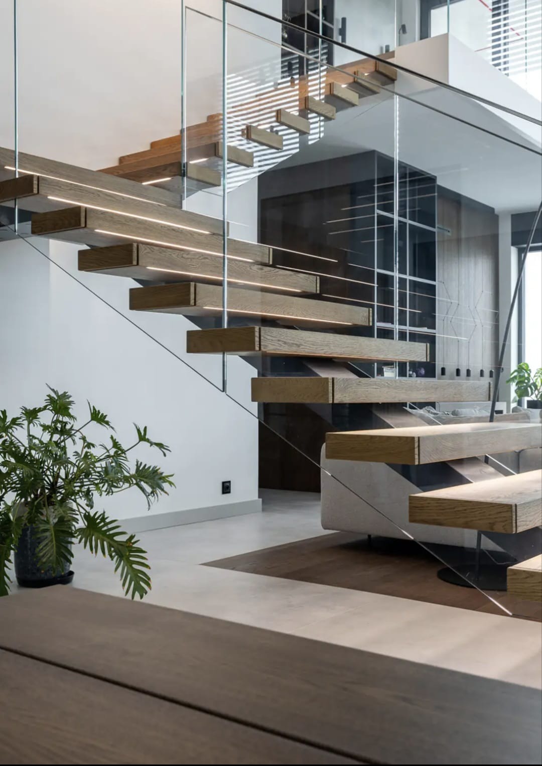 Nowoczesne schody drewniane z podświetleniem LED i szklaną balustradą, widok z dołu, z rośliną doniczkową w tle, minimalistyczny design.