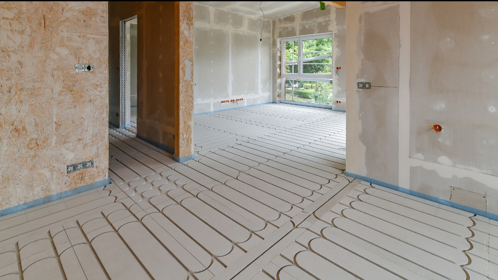 Surowe wnętrze w trakcie prac wykończeniowych: widoczne ogrzewanie podłogowe, ściany przygotowane do malowania, sufit z widoczną instalacją elektryczną, okno z widokiem na zieleń.