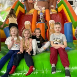 Grupa dzieci siedzi na zielonym, dmuchanym zamku z motywem farmy, w tle widoczna dziewczynka wchodząca na konstrukcję.