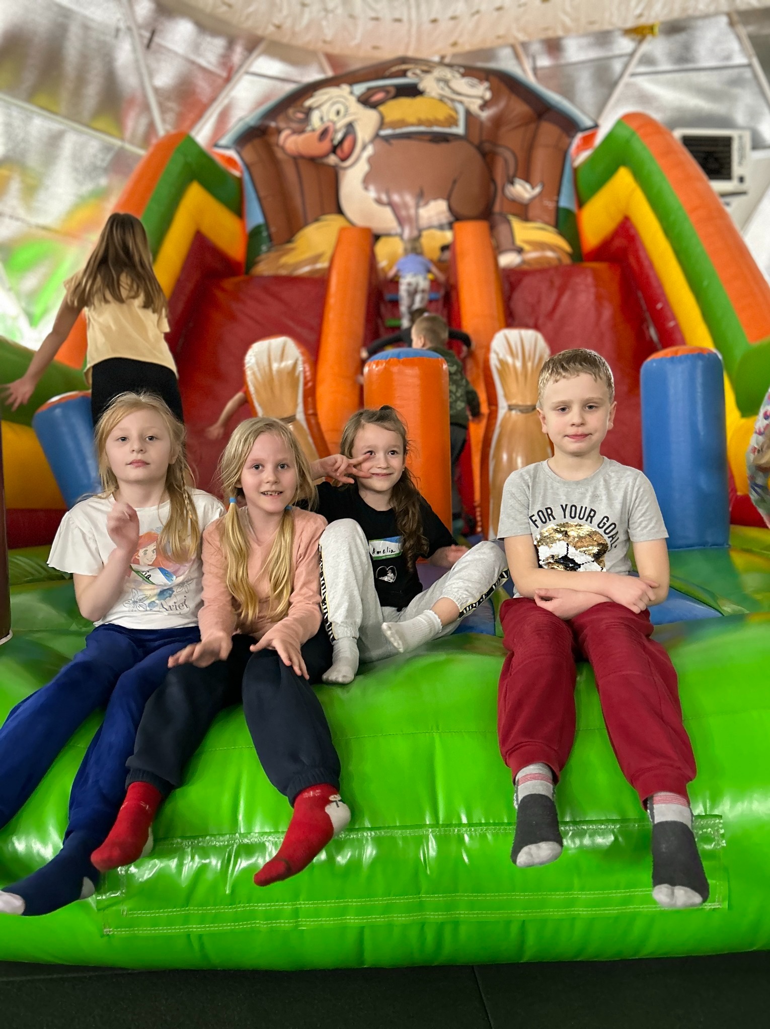 Grupa dzieci siedzi na zielonym, dmuchanym zamku z motywem farmy, w tle widoczna dziewczynka wchodząca na konstrukcję.