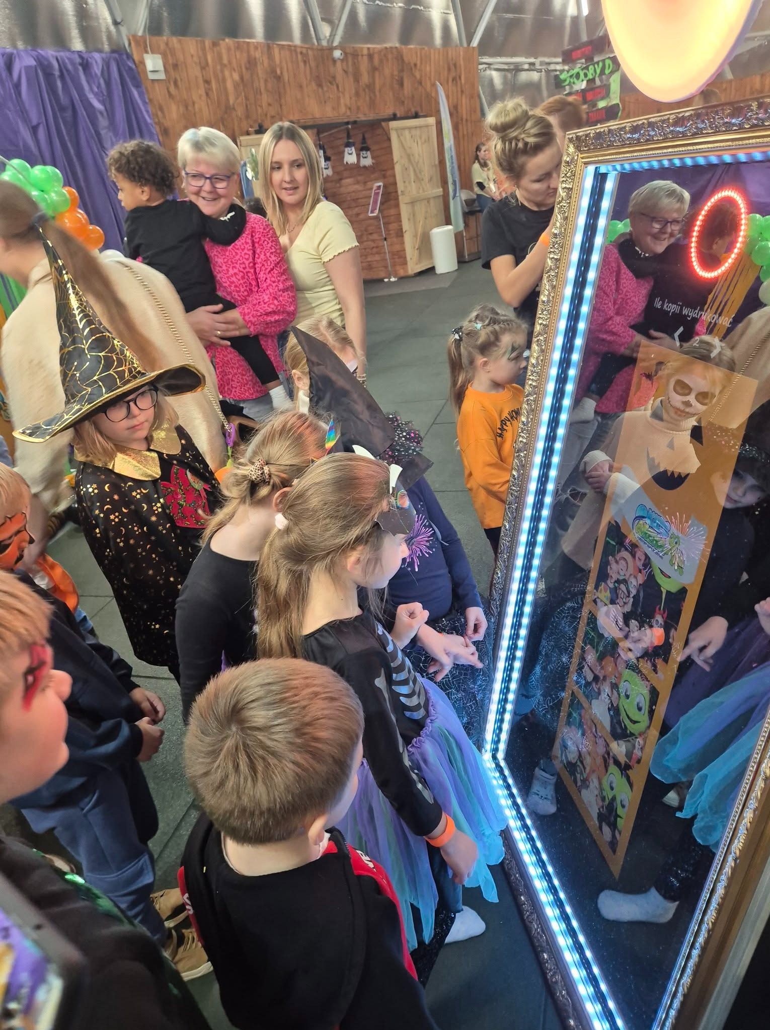 Grupa dzieci w kostiumach halloweenowych pozuje do zdjęcia w fotolustrze z oświetleniem LED i dekoracyjną ramą.