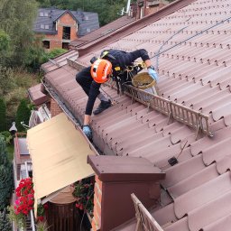 Jakub Nowicki Ruthless Service Usługi Alpinistyczne Prace Wysokościowe - Pracownik w pomarańczowym kasku, zabezpieczony linami, czyści rynnę na stromym, brązowym dachu domu jednorodzinnego, używając do tego rękawic i wiadra z zebranymi zanieczyszczeniami.
