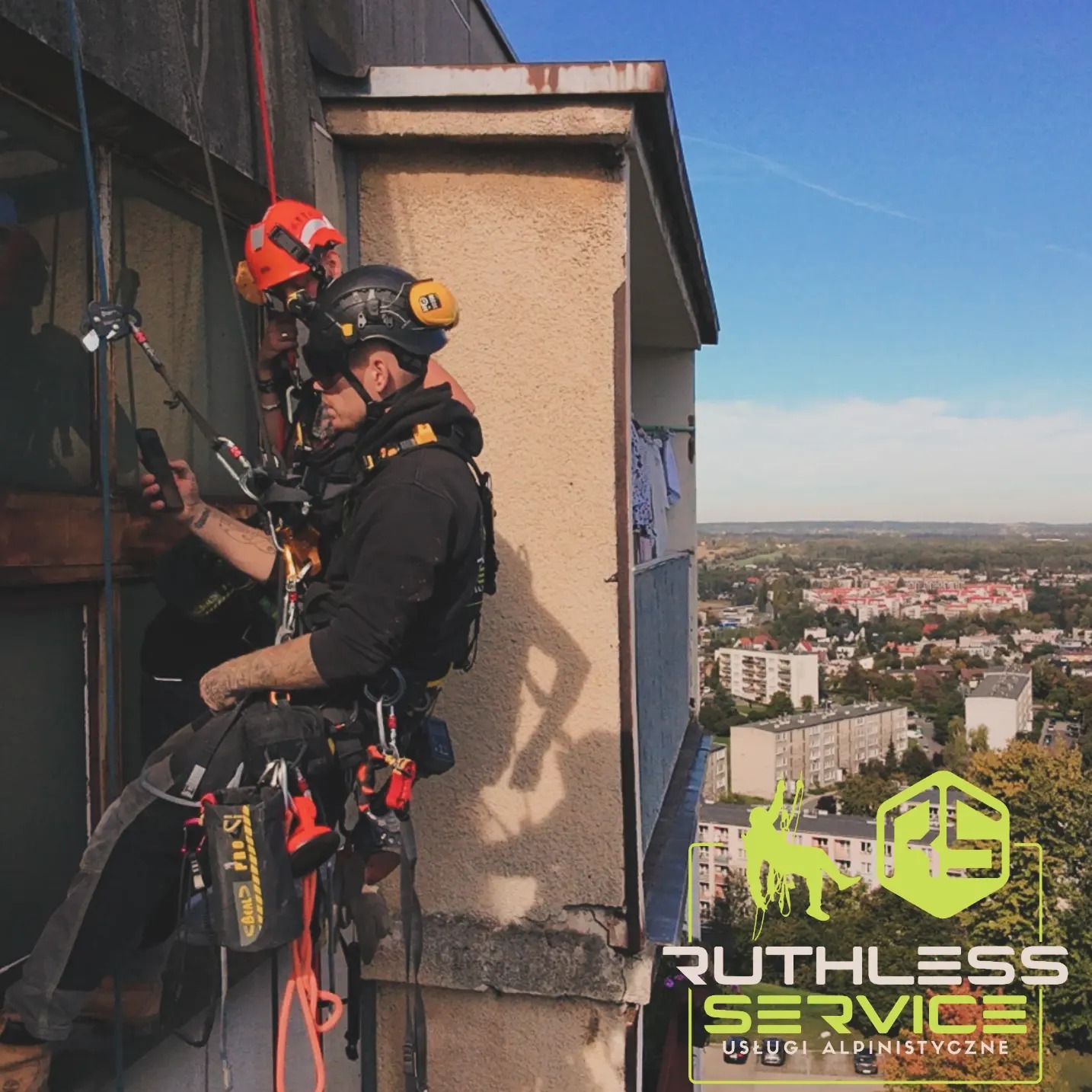 Dwóch alpinistów przemysłowych, jeden z telefonem w dłoni, zawieszonych na linach przy oknie bloku mieszkalnego z widokiem na miasto. W prawym dolnym rogu logo firmy.