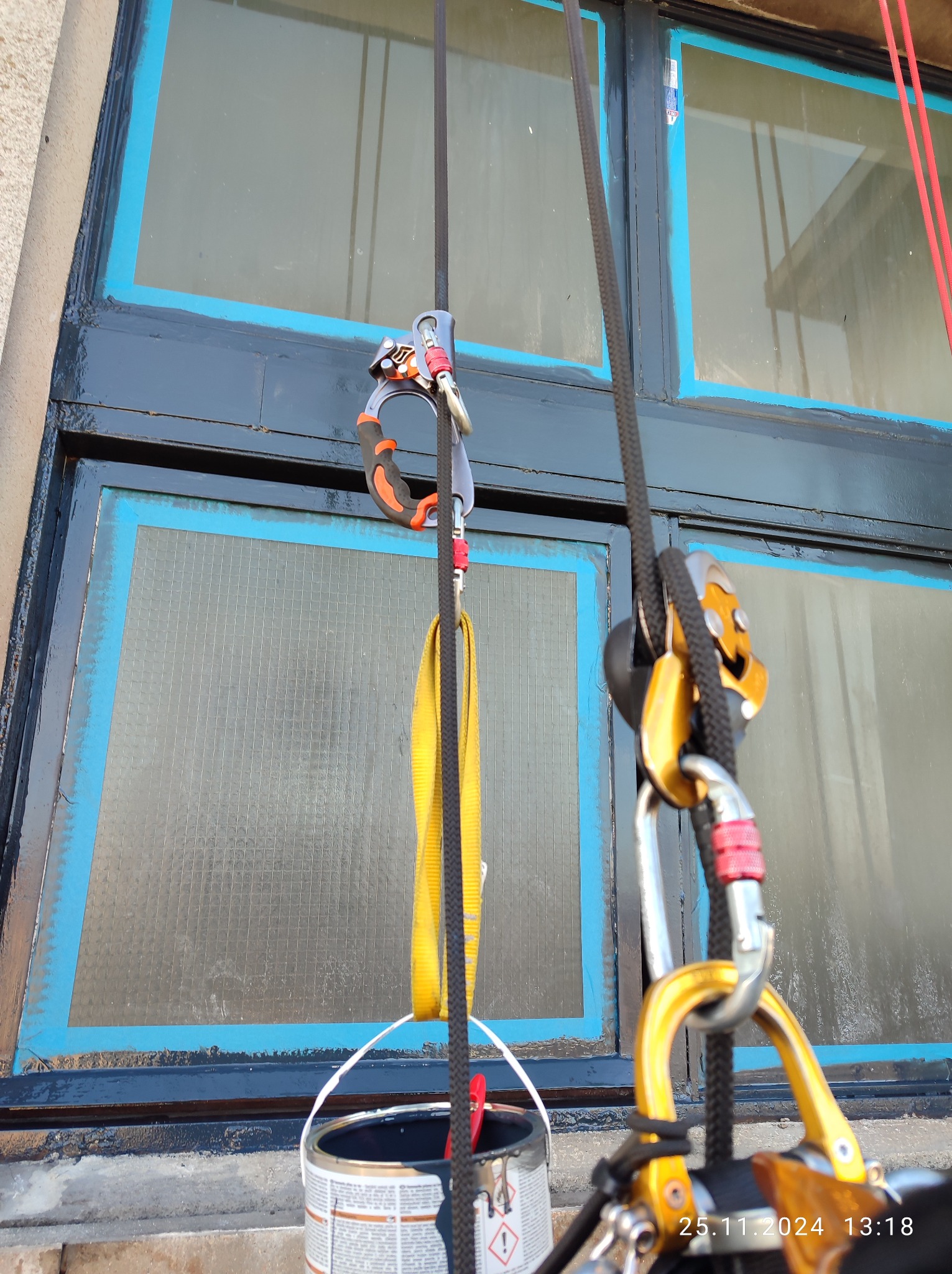 Okna zabezpieczone taśmą malarską, sprzęt do prac wysokościowych na linach, wiadro z farbą widoczne na pierwszym planie, przygotowania do malowania ram okiennych.