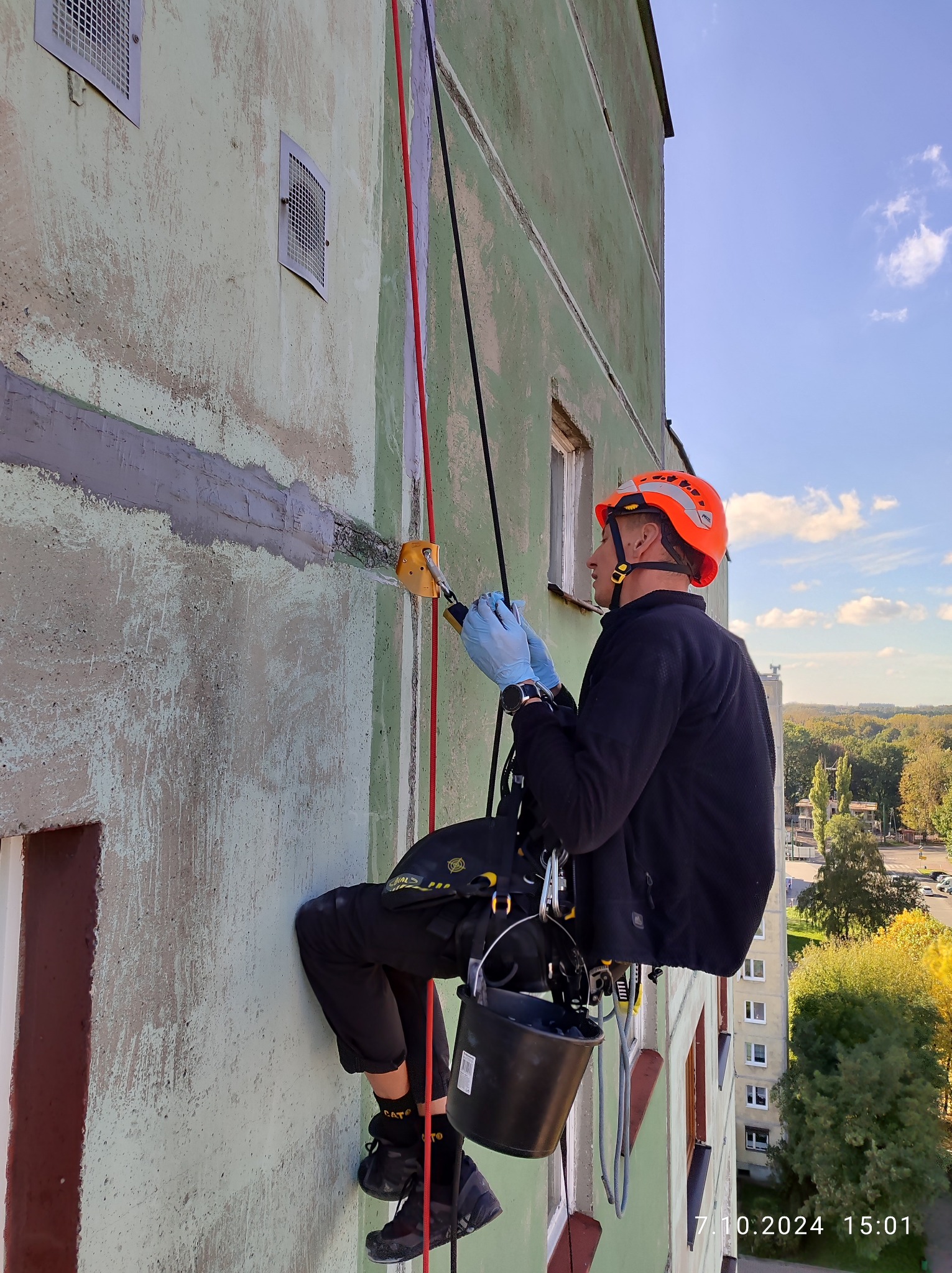 Pracownik w kasku i uprzęży, zawieszony na linach, naprawia uszkodzoną elewację wysokiego budynku w Świętochłowicach, używając narzędzia do wypełniania ubytków.
