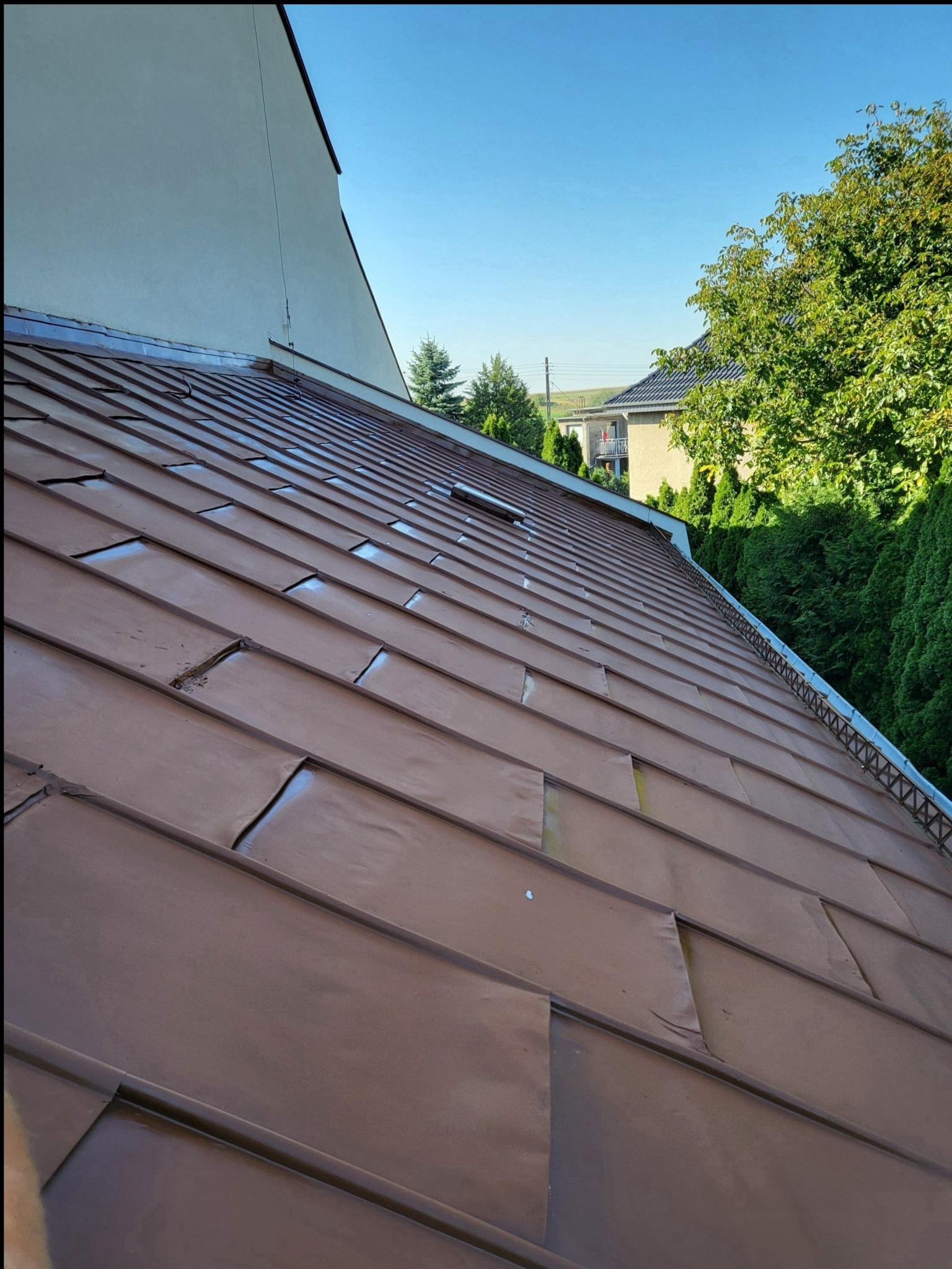 Brązowy dach z blachy trapezowej, widoczne drobne uszkodzenia i zarysowania, w tle budynek mieszkalny i zieleń.
