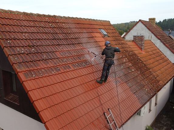 Czyszczenie czerwonej dachówki z mchu myjką ciśnieniową. Widoczna różnica między czystą a zabrudzoną częścią dachu. Pracownik w uprzęży, drabina oparta o budynek.