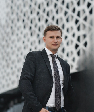 Młody mężczyzna w garniturze i wzorzystym krawacie pozuje na tle nowoczesnej, ażurowej fasady budynku.