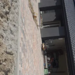 Nowa, wielobarwna kostka ułożona wzdłuż ściany domu z widoczną maszyną do cięcia kostki i przygotowanym terenem pod dalsze prace.