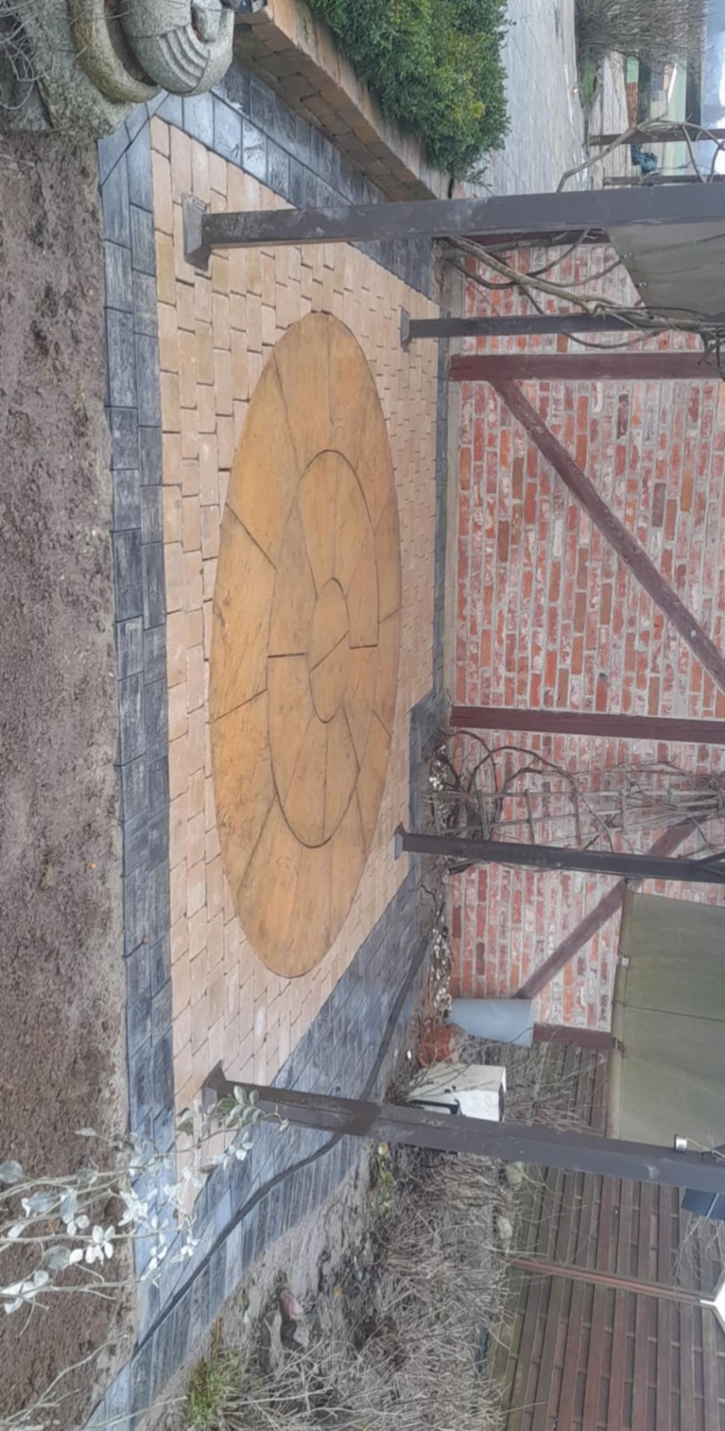 Okrągła kompozycja z beżowej kostki brukowej otoczona ciemnoszarą kostką, tworząca wzór na patio z fragmentem ceglanego muru i metalowej konstrukcji zadaszenia w tle.