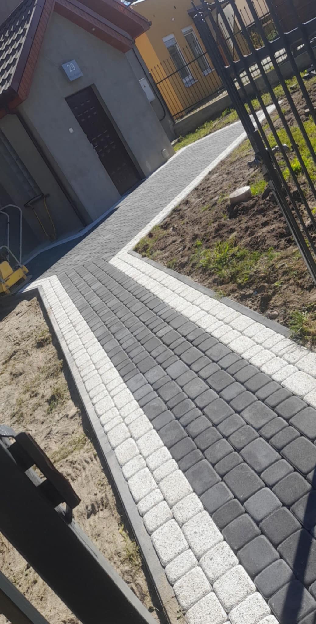 Ścieżka z szarej i białej kostki brukowej prowadząca do drzwi budynku z numerem 29, widok z poziomu gruntu, częściowo widoczne ogrodzenie metalowe.