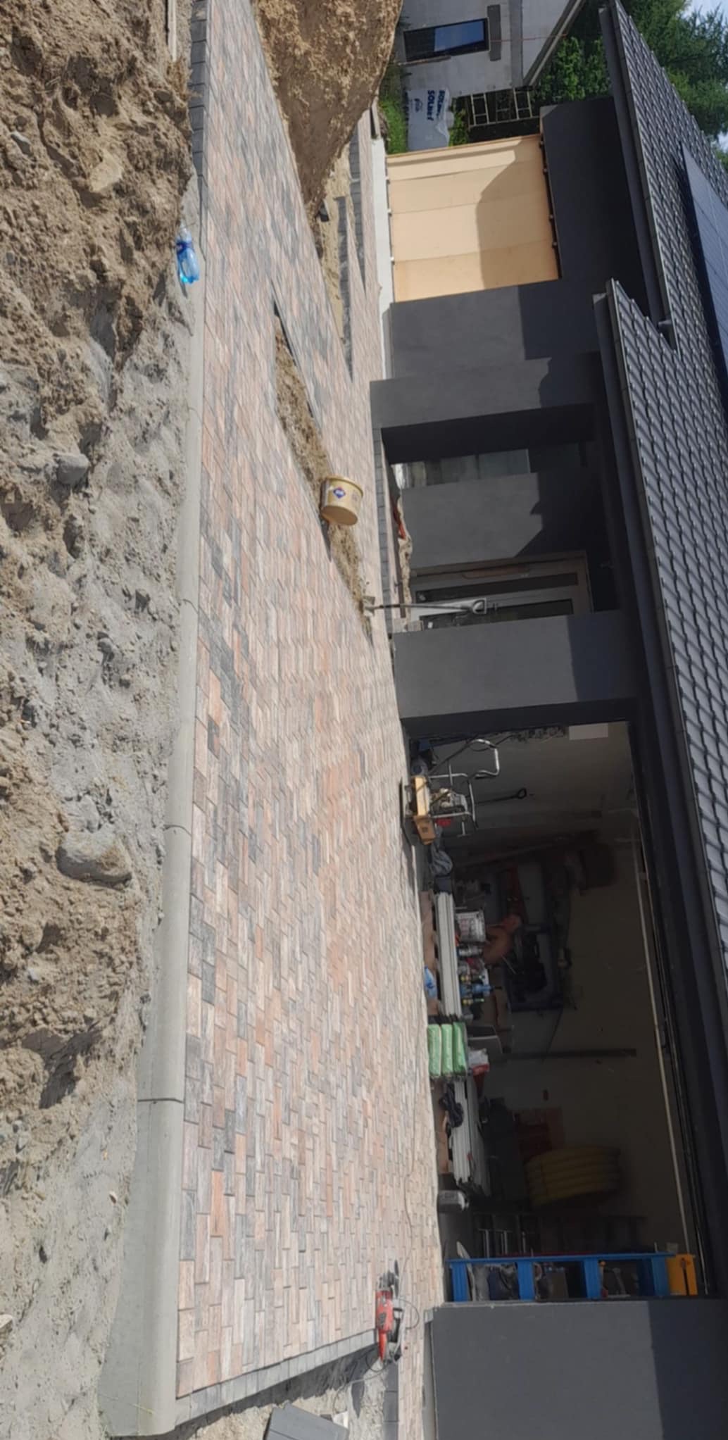 Nowa, wielobarwna kostka ułożona wzdłuż ściany domu z widoczną maszyną do cięcia kostki i przygotowanym terenem pod dalsze prace.