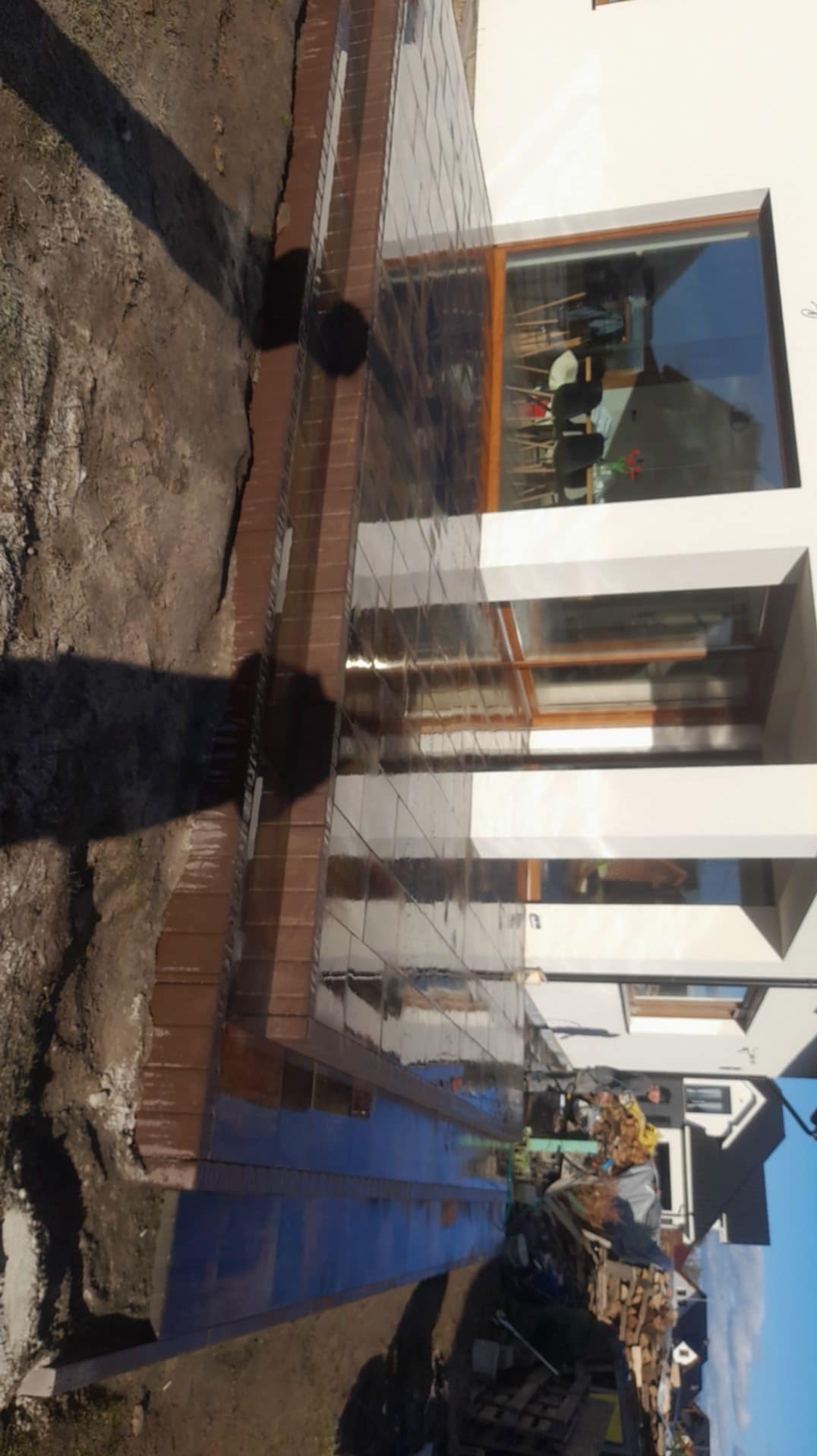 Nowo ułożona, błyszcząca, ciemna kostka brukowa przed nowoczesnym domem z dużym oknem, widoczne ślady po pracy i stos drewna w tle.