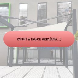 Kasia Skrobotowicz - Zadaszenie tarasowe z białym żaglem przeciwsłonecznym, metalowa konstrukcja na tle budynku z szarymi elewacjami i zielonymi ramami okiennymi, widoczny fragment chodnika z jasnej kostki brukowej.