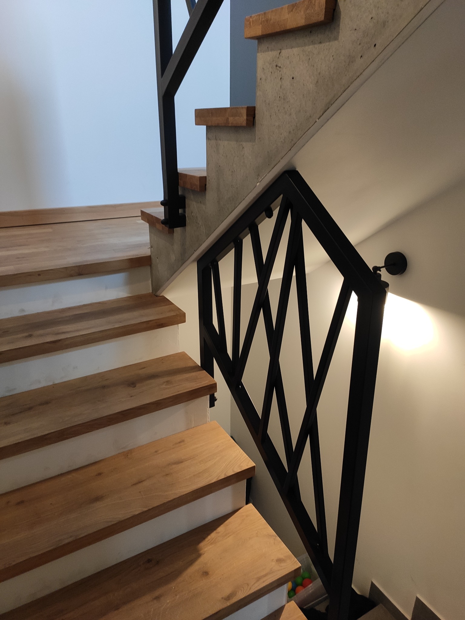 Nowoczesne schody z drewnianymi stopniami i czarną, geometryczną balustradą. Ściany pomalowane na biało, oświetlenie punktowe.