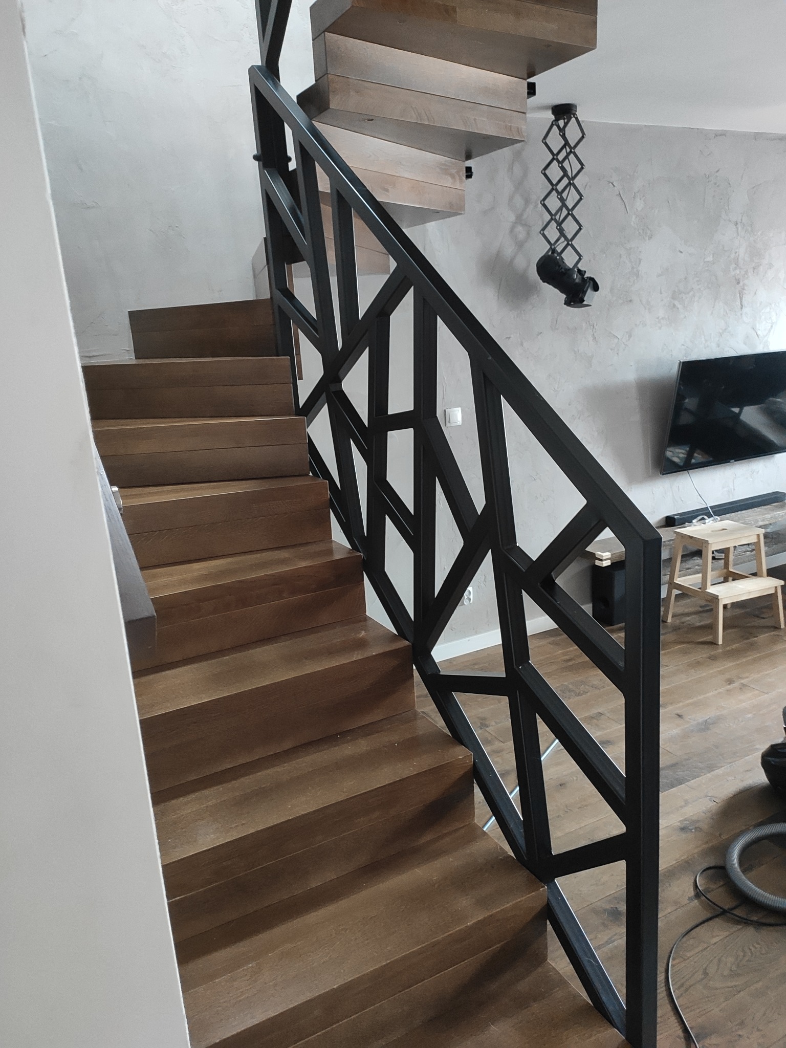 Drewniane schody z geometryczną, czarną, metalową balustradą. Na ścianie widoczna lampa sufitowa w stylu industrialnym oraz telewizor. Pod schodami widoczny odkurzacz.