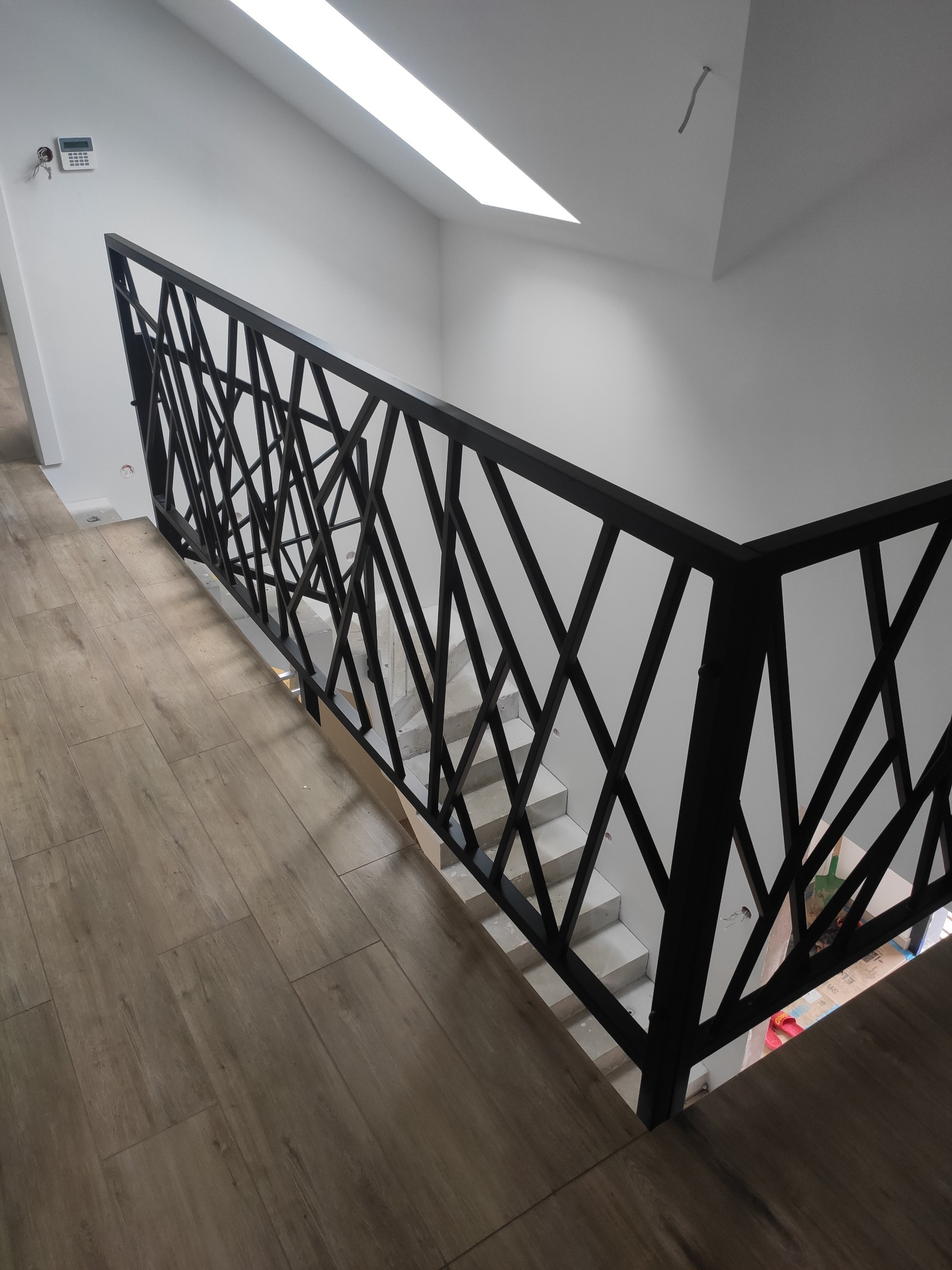 Nowoczesna, czarna barierka z geometrycznym wzorem na białych schodach, widziana z góry, z fragmentem drewnopodobnej podłogi i białymi ścianami w tle.