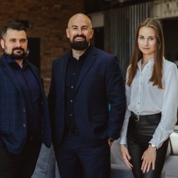 FENBRO SPÓŁKA Z OGRANICZONĄ ODPOWIEDZIALNOŚCIĄ - Portret zespołu: mężczyzna w kraciastej marynarce, łysy mężczyzna z brodą w granatowym garniturze i kobieta w białej koszuli i skórzanych spodniach, stojący w nowoczesnym biurze z ceglaną ścianą...